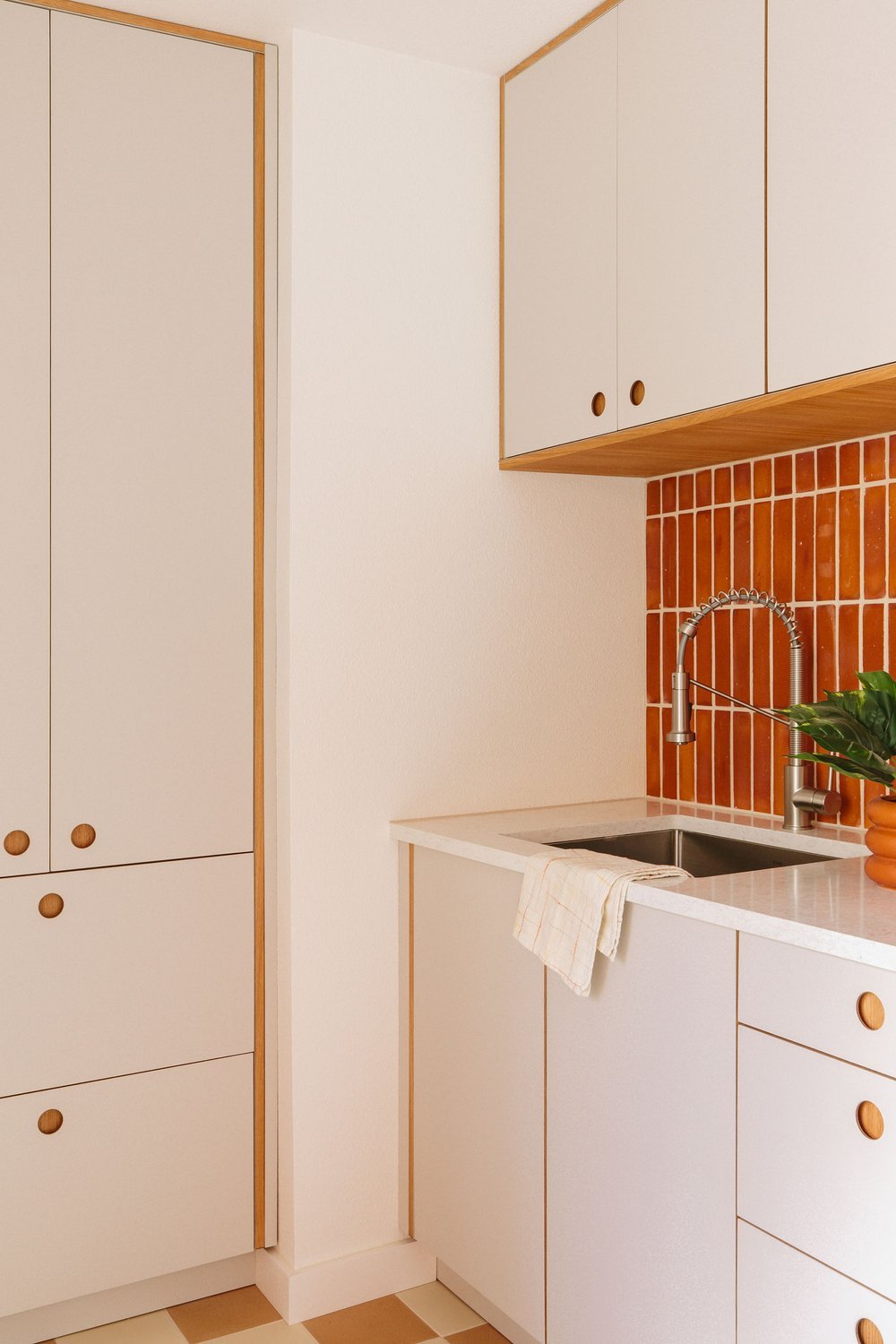Minimalist kitchen with white cabinets, a red tiled backsplash, a stainless steel sink with a modern faucet, and a potted green plant on the counter.