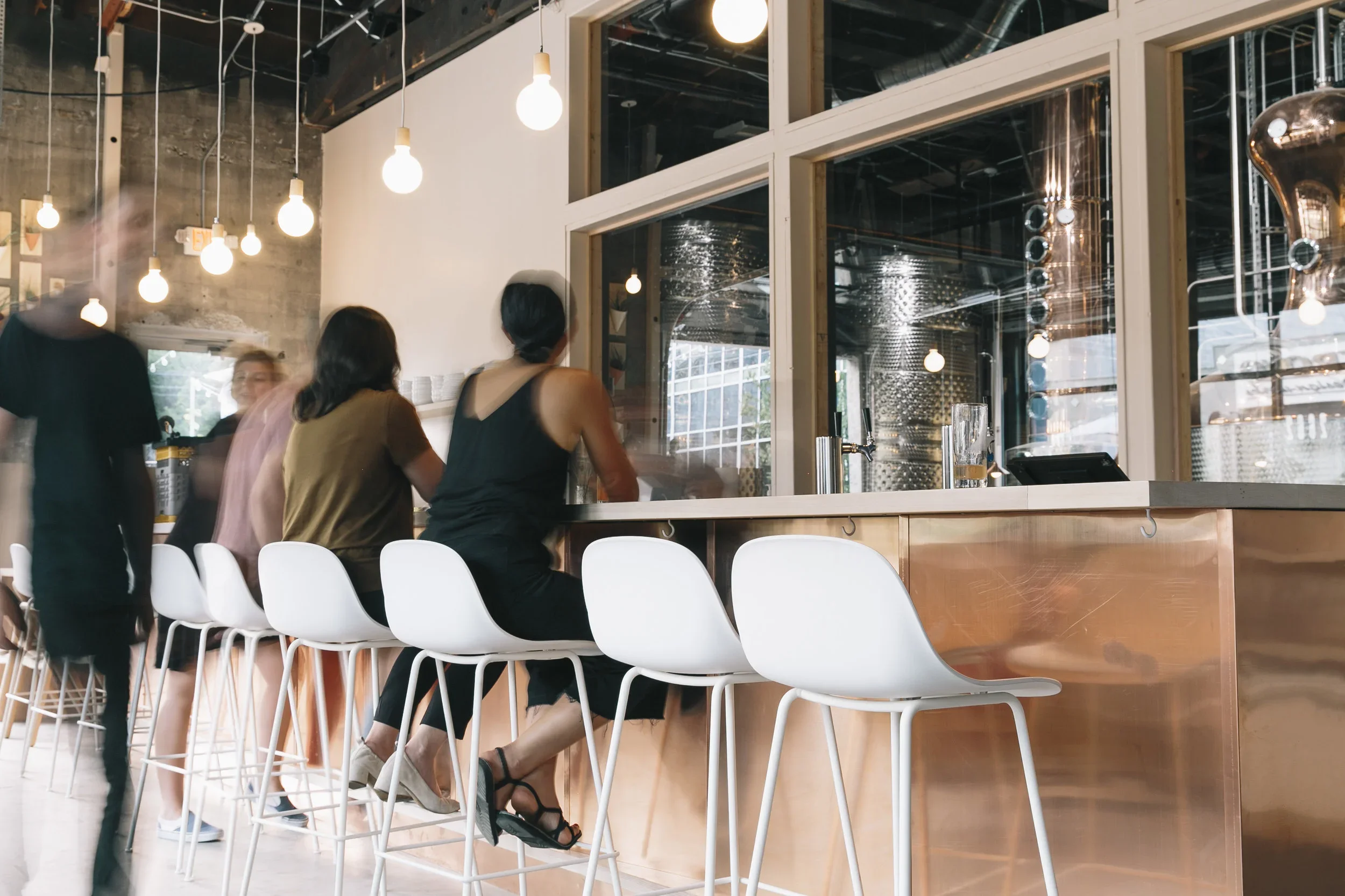People sitting at a bar counter in a modern, stylish café or restaurant with hanging light bulbs and industrial decor.