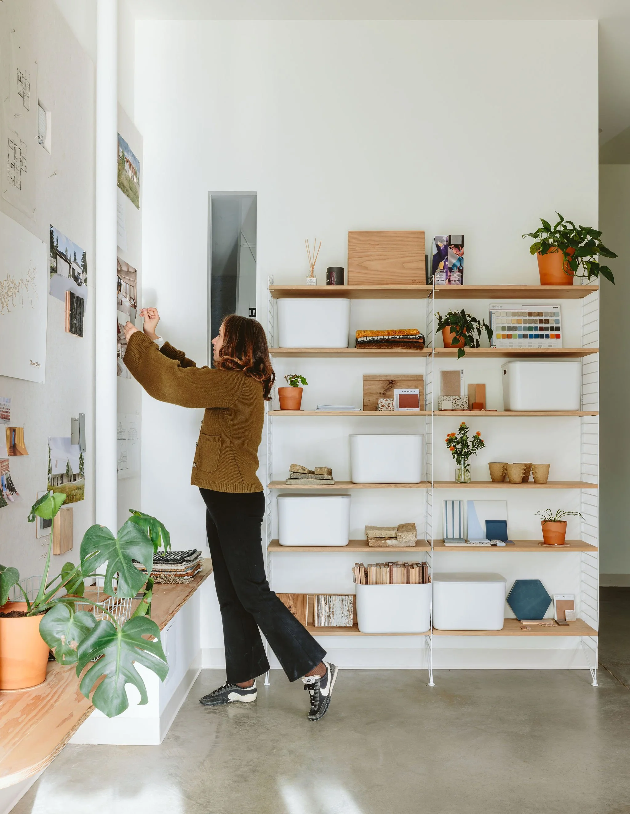 A woman with brown hair, wearing a brown sweater and black pants, is sticking photos or artwork to a white wall in a room with a bookshelf, potted plants, and decorative items.