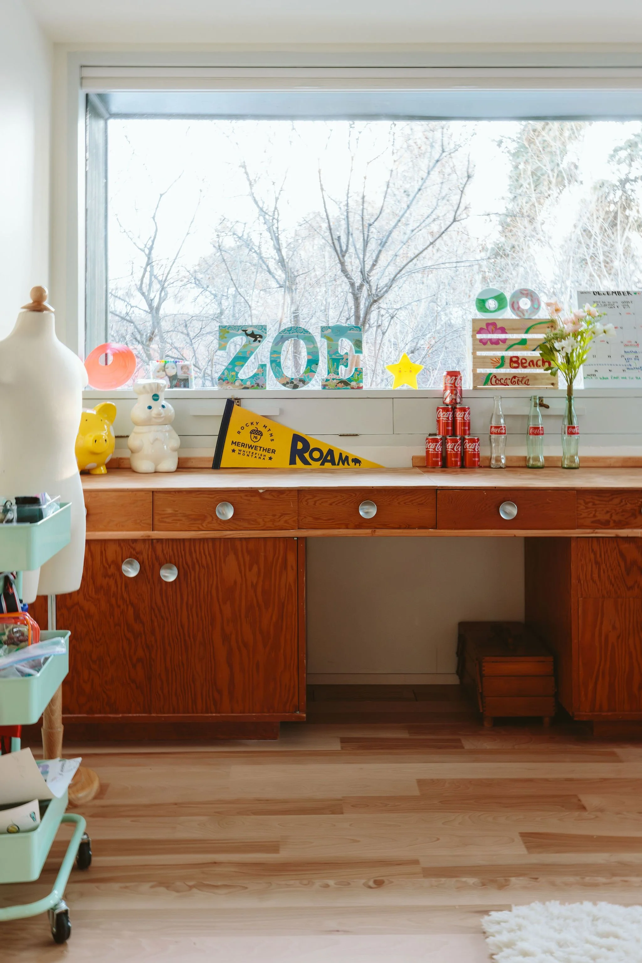 Decorated wooden desk in front of a large window with snow-covered trees outside, featuring colorful signs, Coca-Cola cans, glass bottles, flowers, and small figurines.