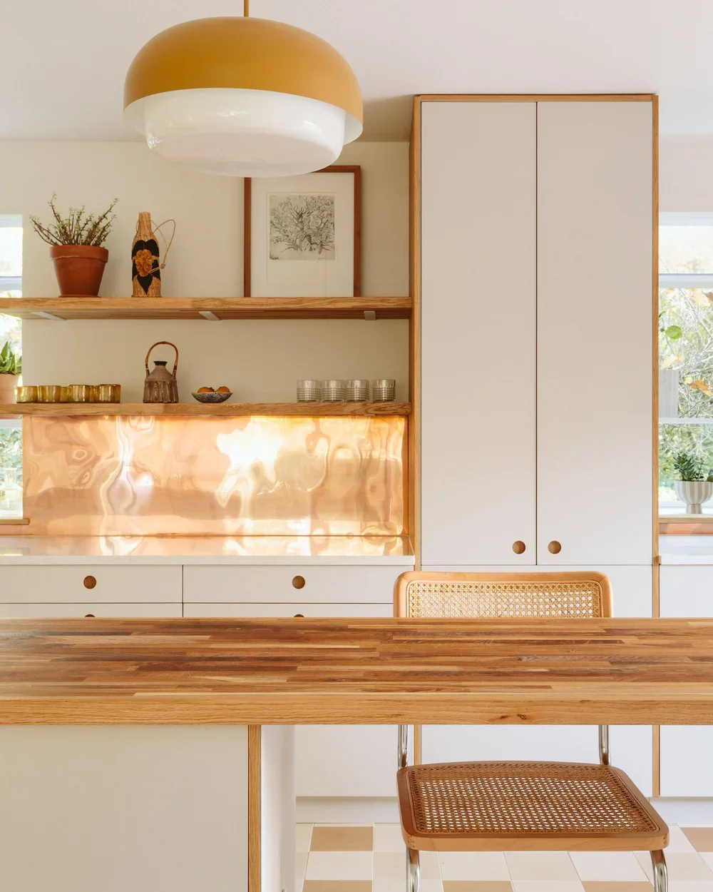 A modern kitchen with wooden shelves, white cabinetry, a copper backsplash, and a wooden table with rattan chair.