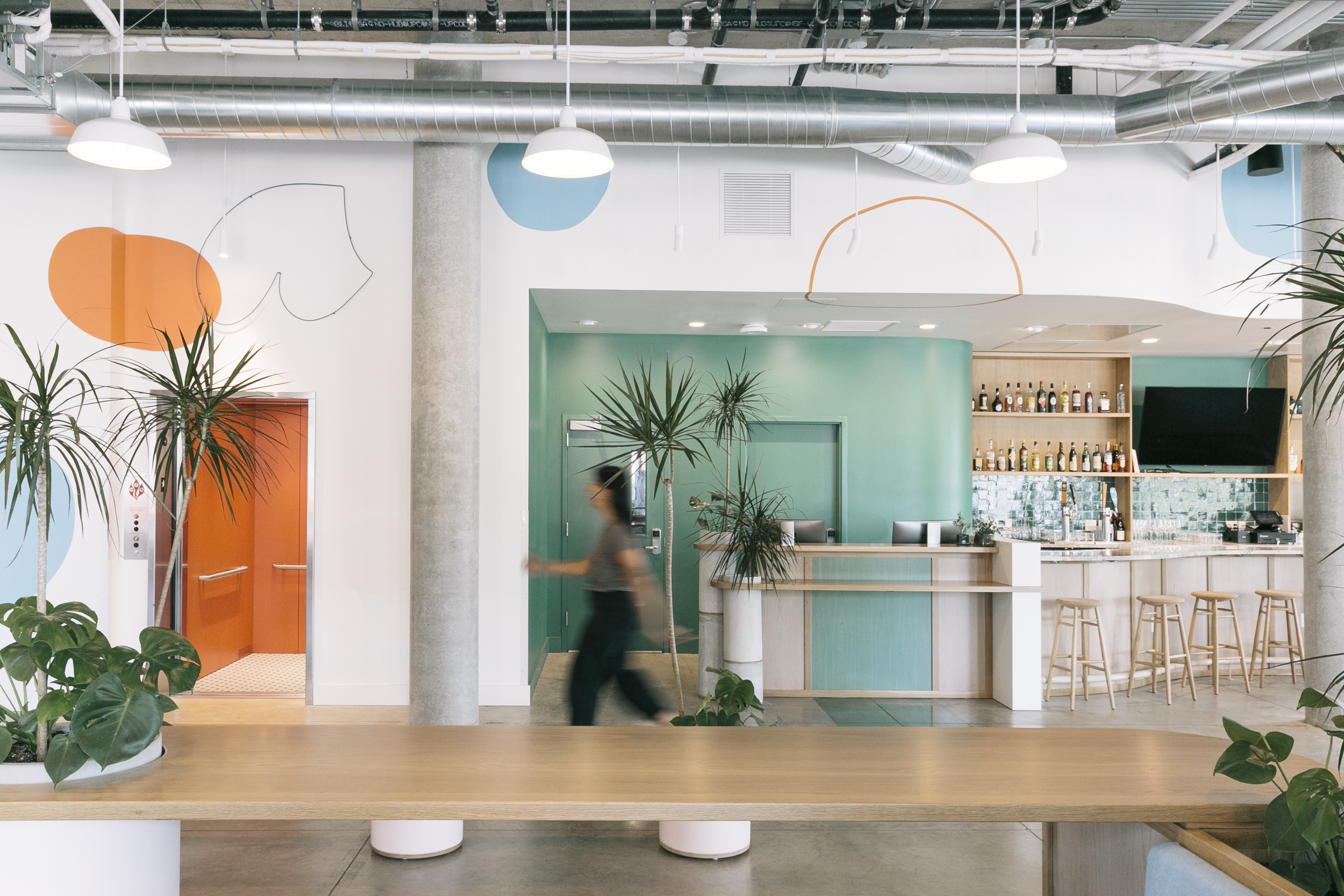Modern, colorful cafe interior with green accent wall, bar area with stools, plants, and a person walking past."}