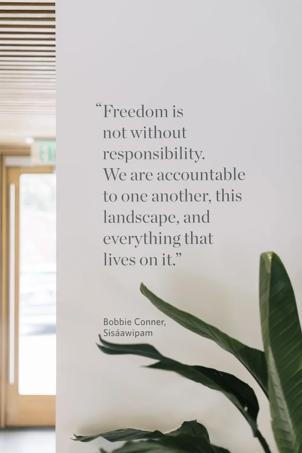 A quote on a white wall that reads, "Freedom is not without responsibility. We are accountable to one another, this landscape, and everything that lives on it." by Bobbie Conner, Sisāwipam. There is a green plant in the foreground and a doorway with an exit sign in the background.