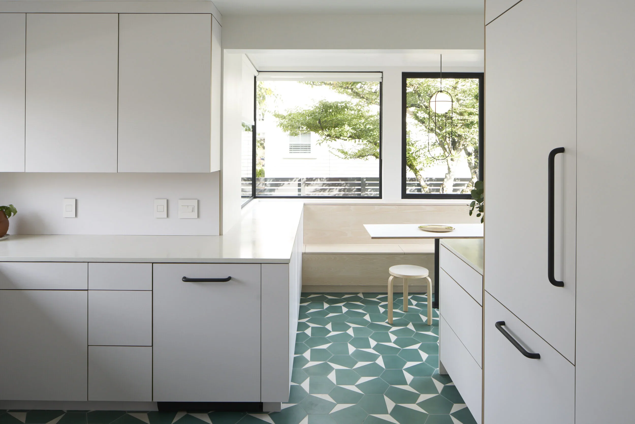 Modern kitchen with white cabinets, geometric patterned teal and white floor tiles, large windows, and a small seating area with a wooden stool.