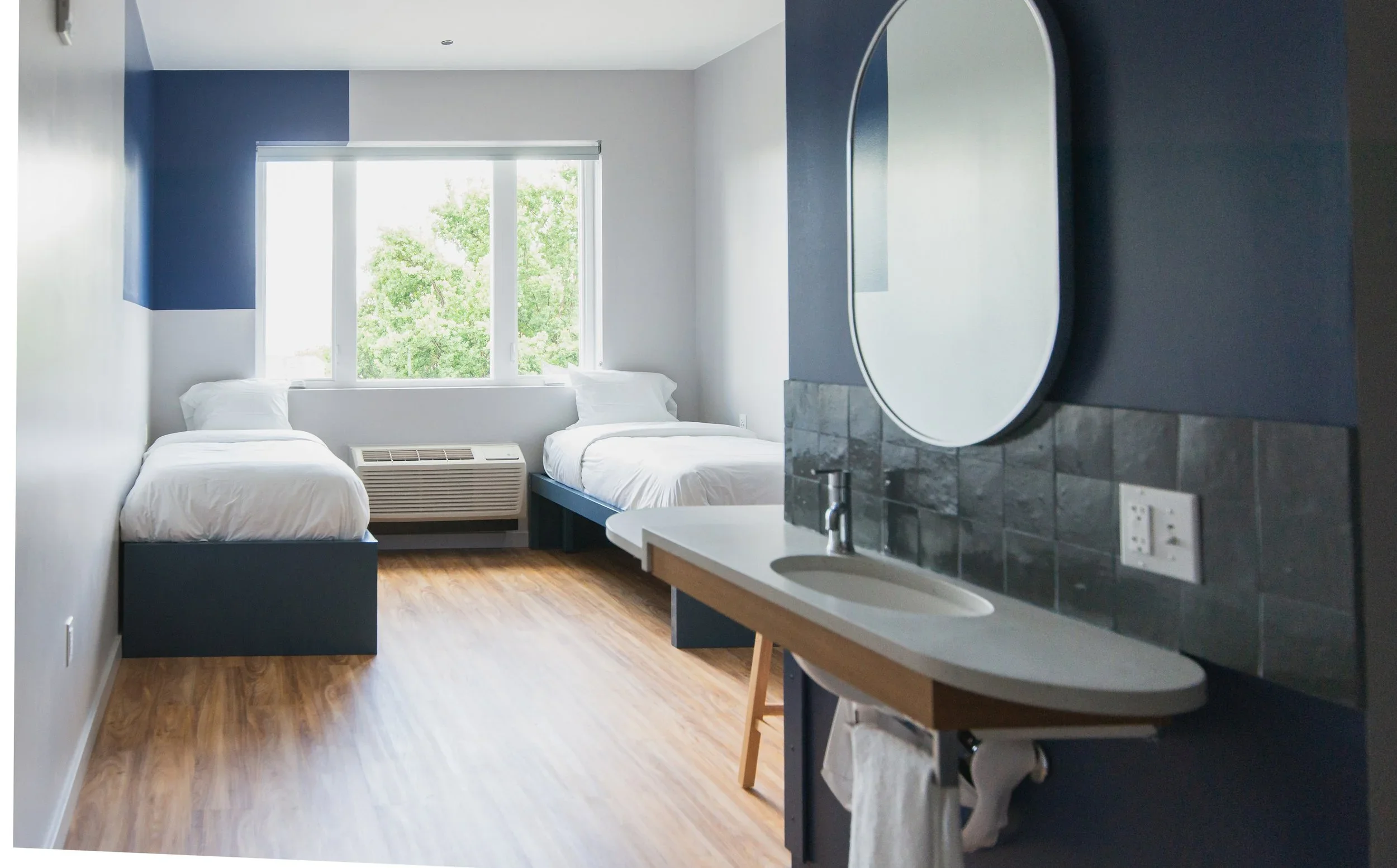 Interior of a modern hotel room with two beds, a window with green trees outside, a sink with a mirror, and hardwood floors.