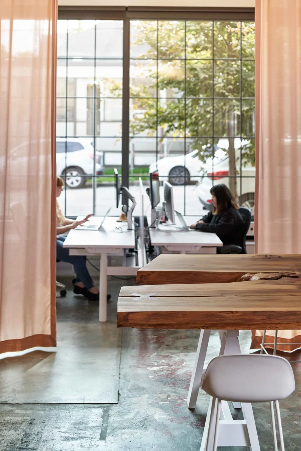 View through a window showing two women working at desks with computers, separated by curtains in a modern office with concrete floors and large glass windows.