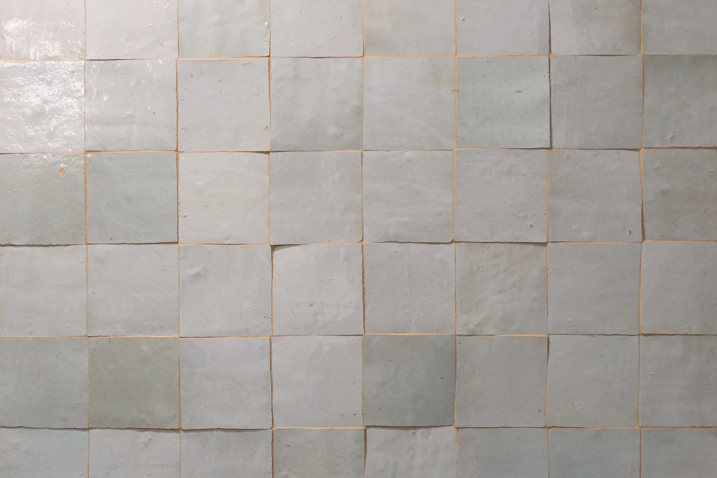 A wall of square ceramic tiles in a grid pattern, mostly light gray, with some tiles slightly darker or with minor imperfections.