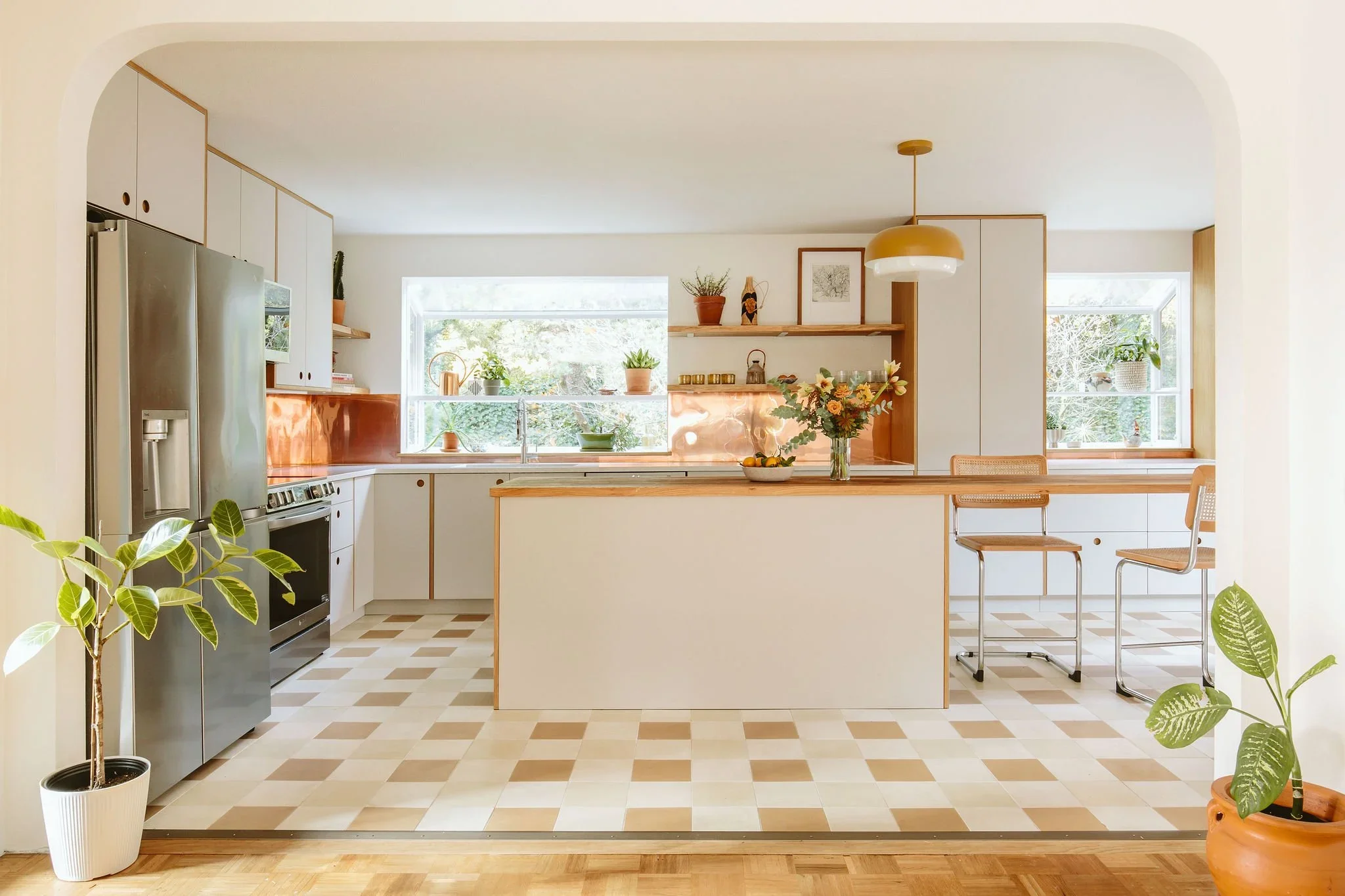 Bright kitchen with white cabinets, a wooden kitchen island, stainless steel refrigerator, and two bar stools. Large windows bring in natural light, decorated with potted plants and flowers. Light-colored tile flooring and minimalistic decor create a cozy atmosphere.
