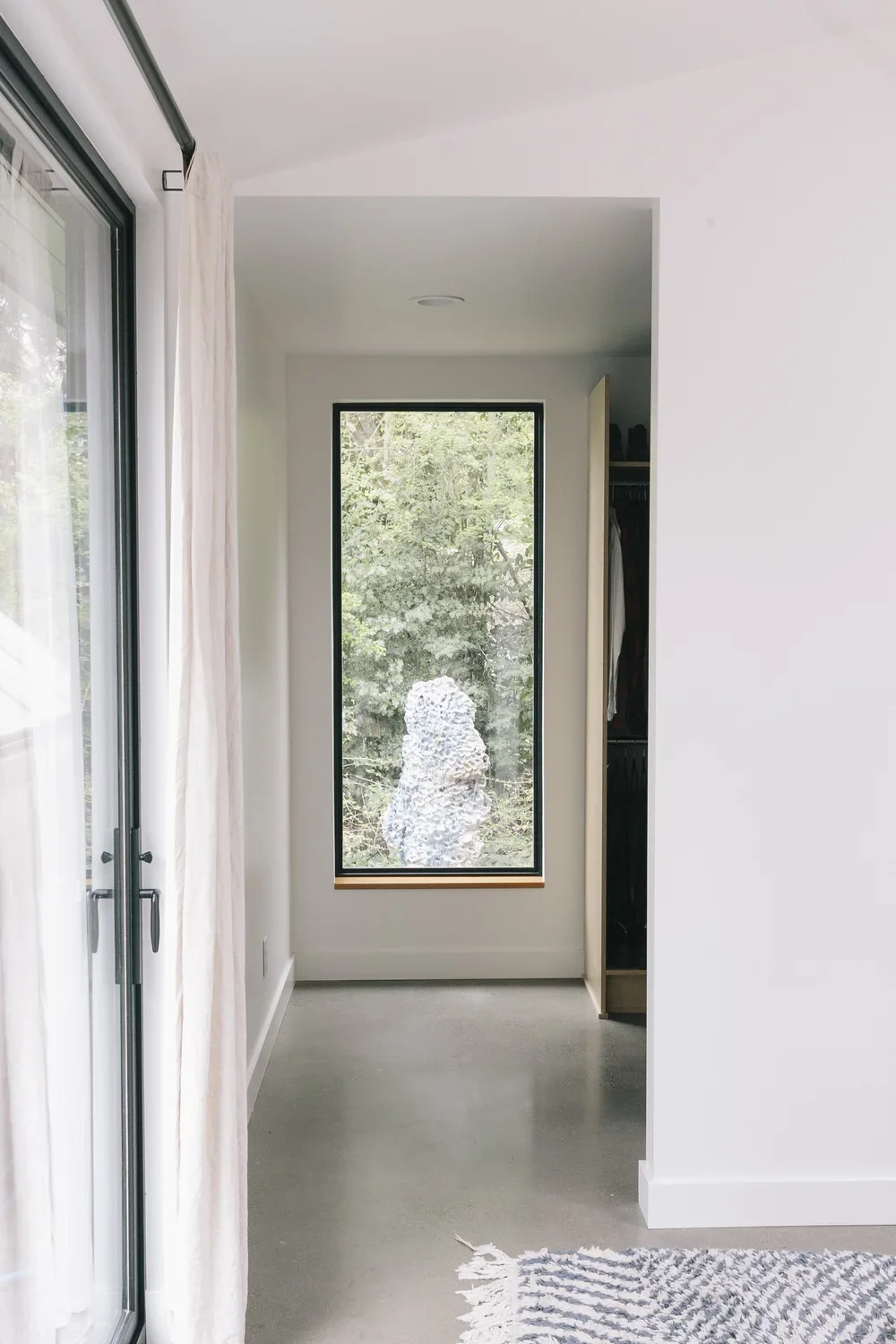 Modern interior space with a large window showing a textured sculpture outside, white walls, a closet with hanging clothes, and a doorway with curtains.