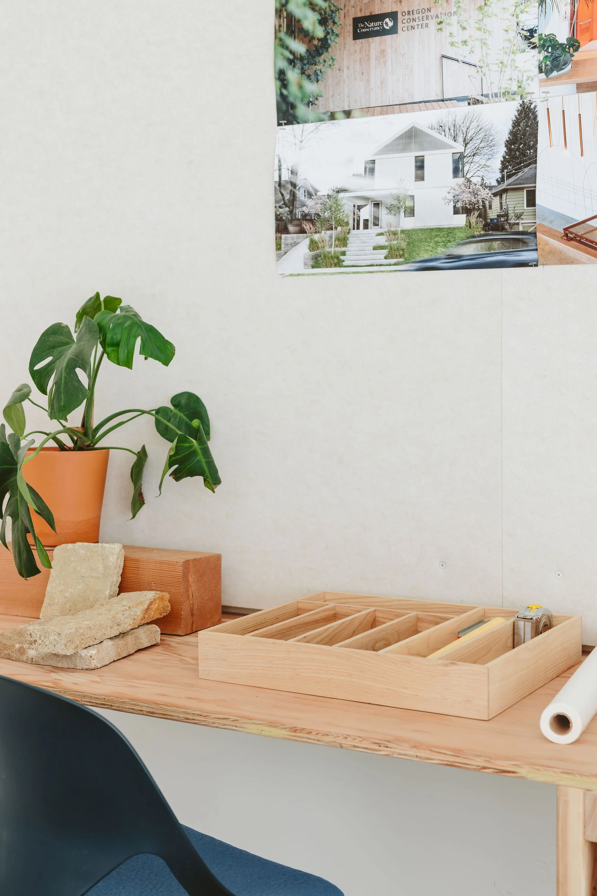 Desk with a potted plant, a wooden box organizer, a roll of paper, and construction materials on it, with photos of houses and a wooden sign in the background.
