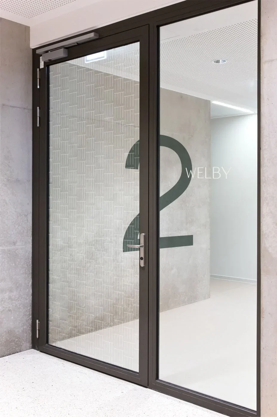 Glass door with black frame, door handle, and the number 2 with the name Welby on the glass, leading into a modern interior space with light-colored walls and ceiling.