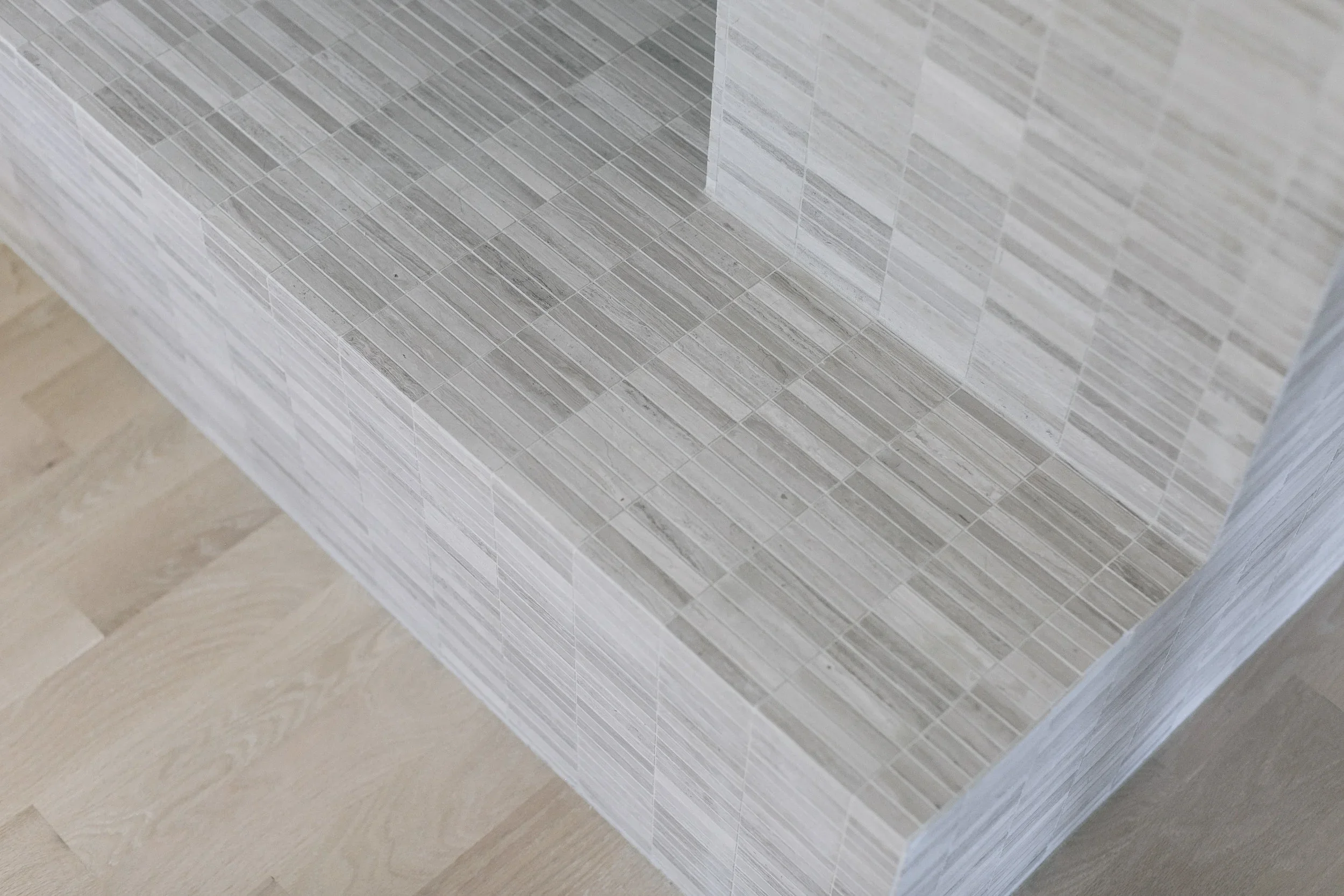 Close-up view of a tiled corner wall with beige and gray rectangular tiles in a diagonal pattern, and a wooden floor at the base.