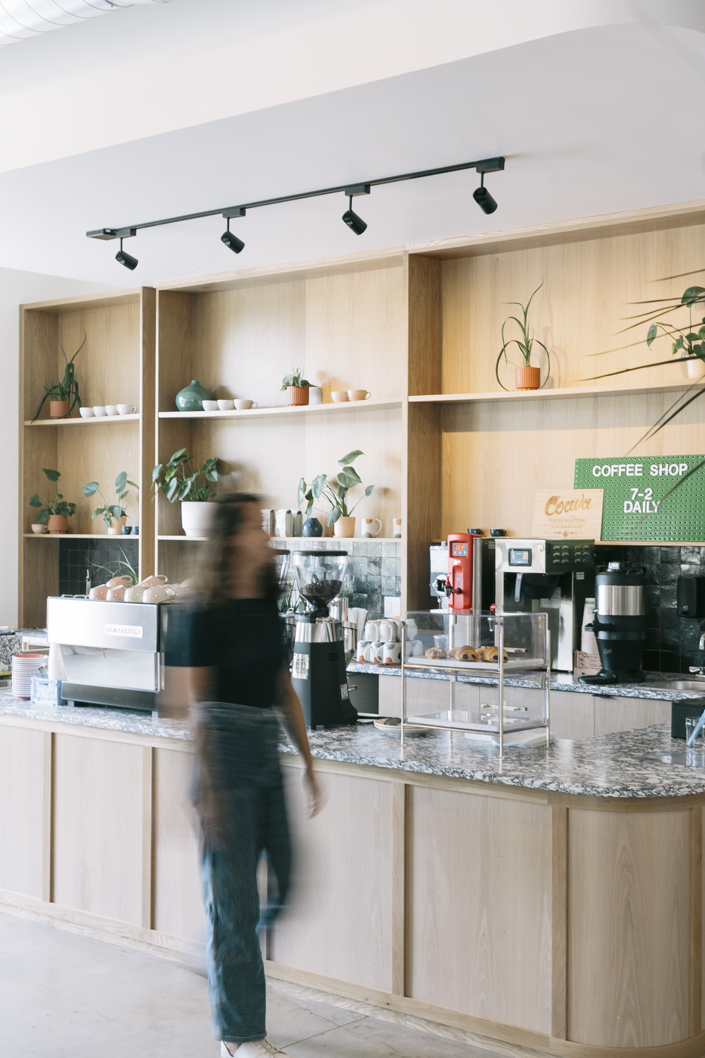 Coffee shop interior with a light wood counter, coffee machines, pastries, and shelves with potted plants and cups, with a woman walking past in motion blur.