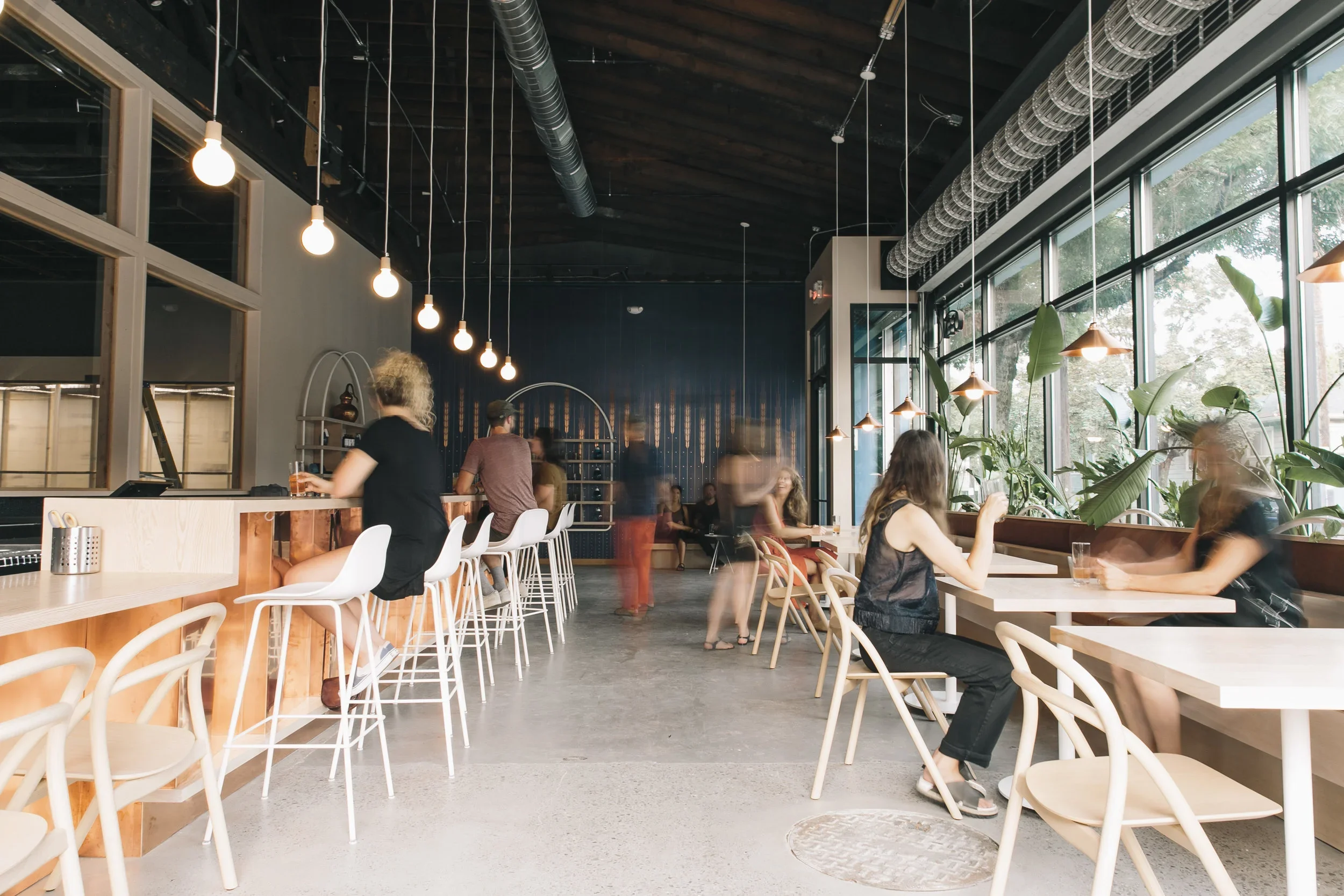 Interior of a modern cafe with high ceiling, hanging lights, large windows, and multiple people sitting and standing in the space.