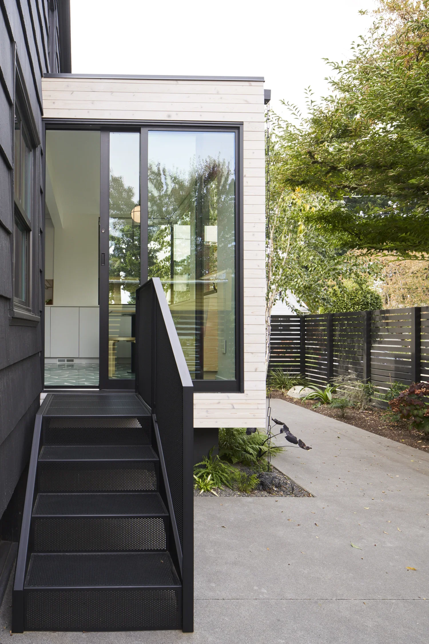 Exterior view of a modern house with black steps leading to a glass sliding door, a small garden with greenery, and a black wooden fence.