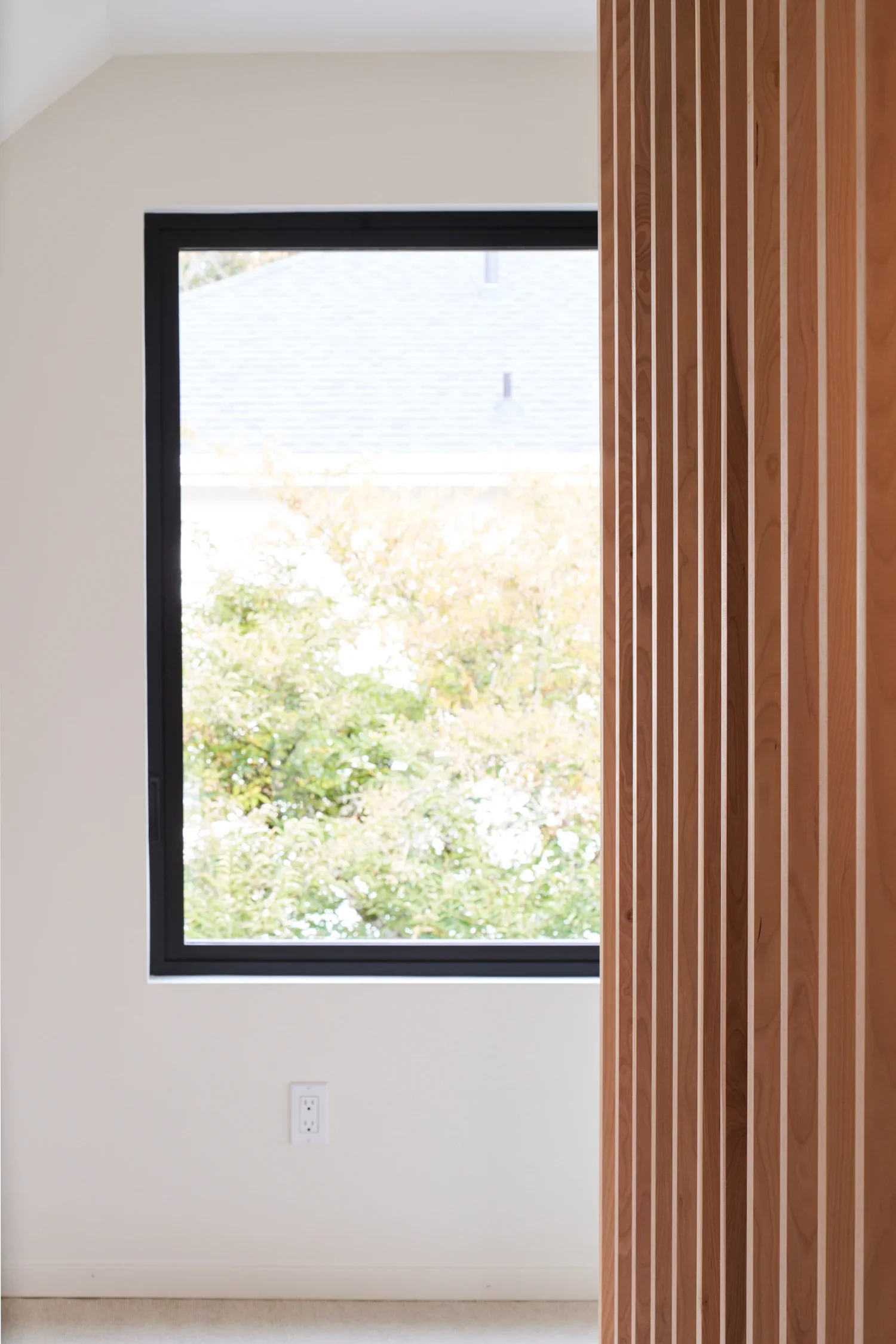 Interior view of a modern room with a window showing outside trees and roof, light-colored wall with a black window frame, and a partial view of a wooden slat wall or panel.