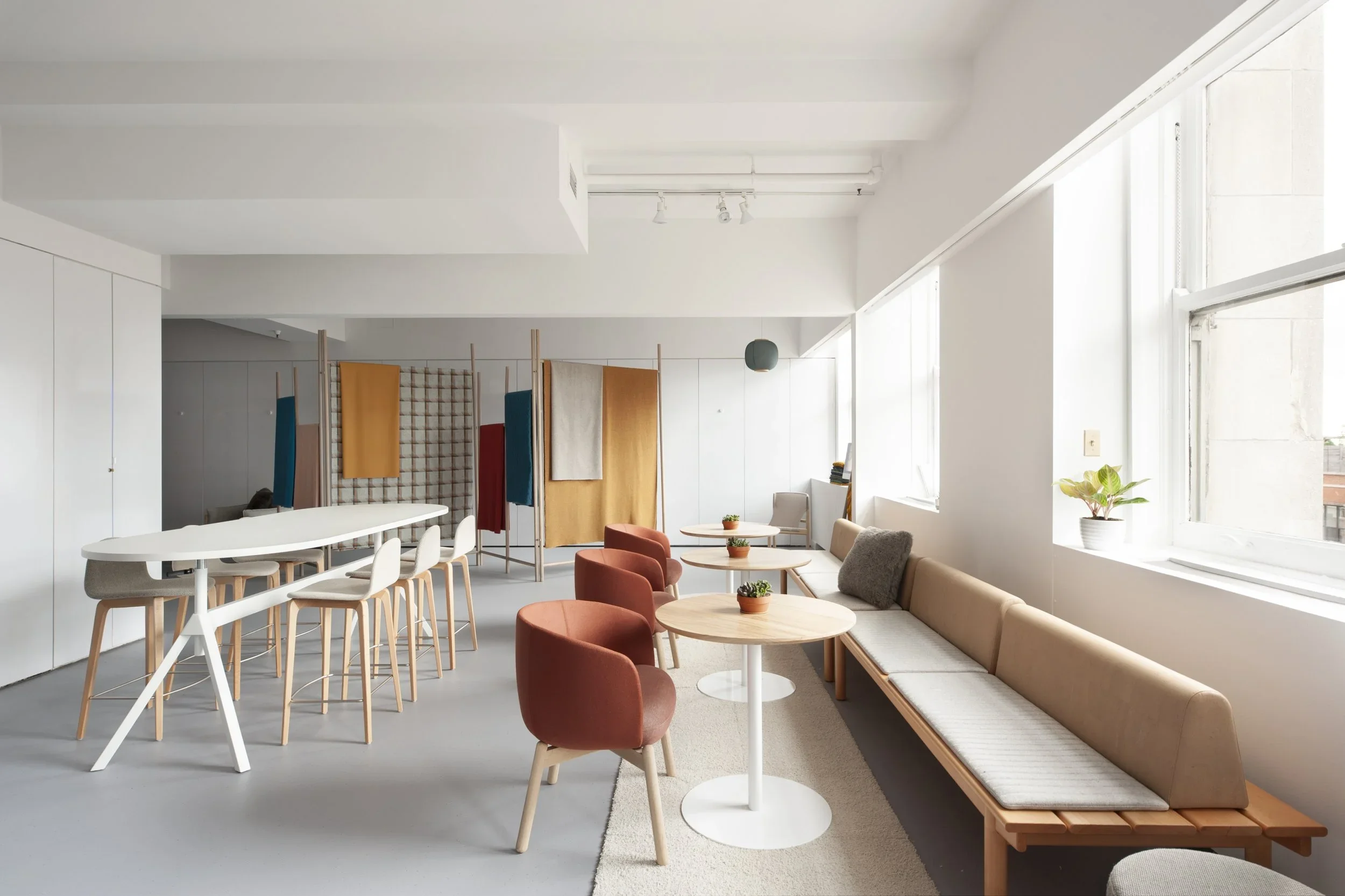 A well-lit modern lounge or workspace with beige and rust-colored chairs, small round tables, a long beige bench with gray cushions, and colorful fabric dividers in the background, all set against large windows and white walls.