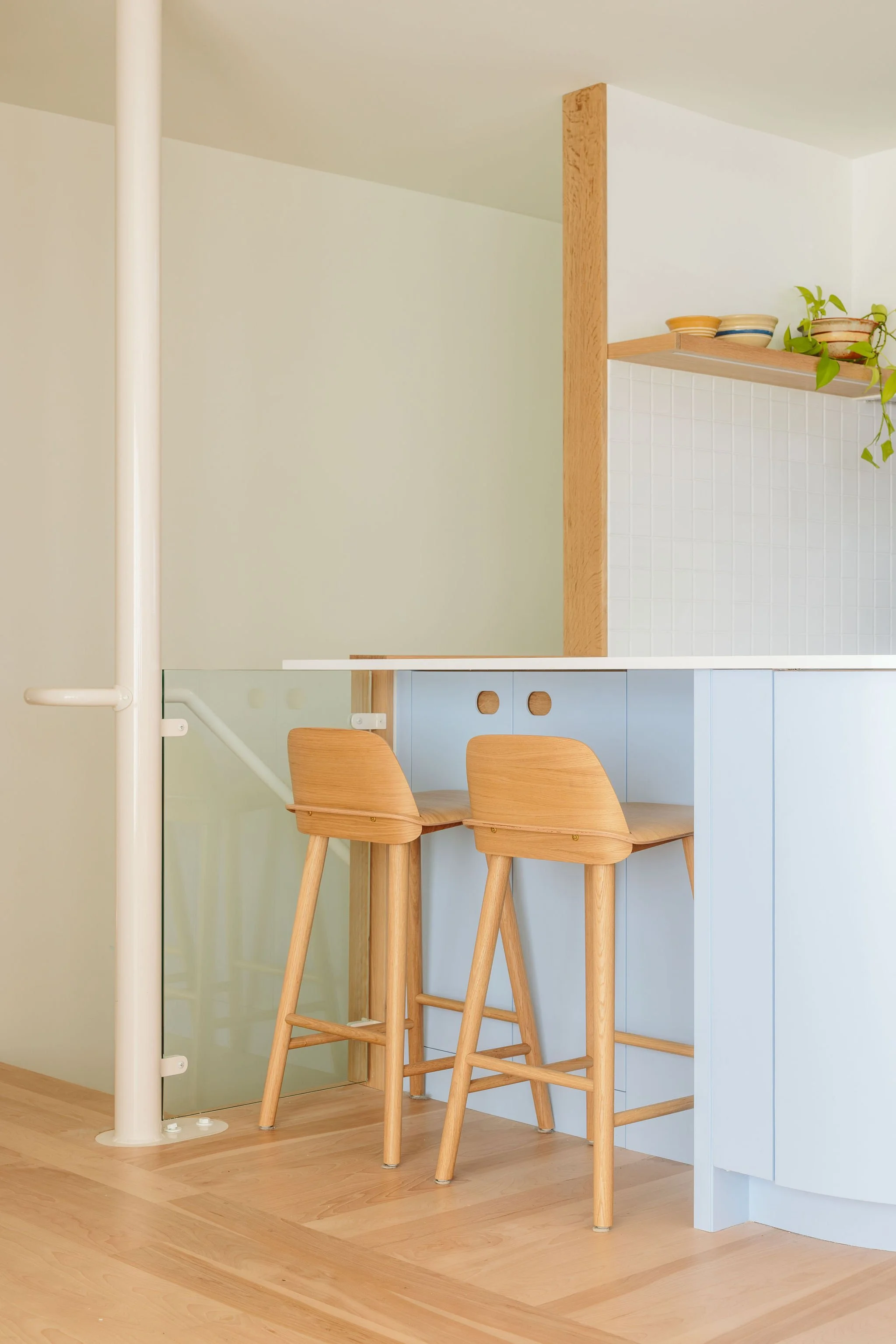 A minimalist kitchen nook with two wooden bar stools at a white counter, partially enclosed by a glass and white panel, with a shelf holding bowls and a plant above.