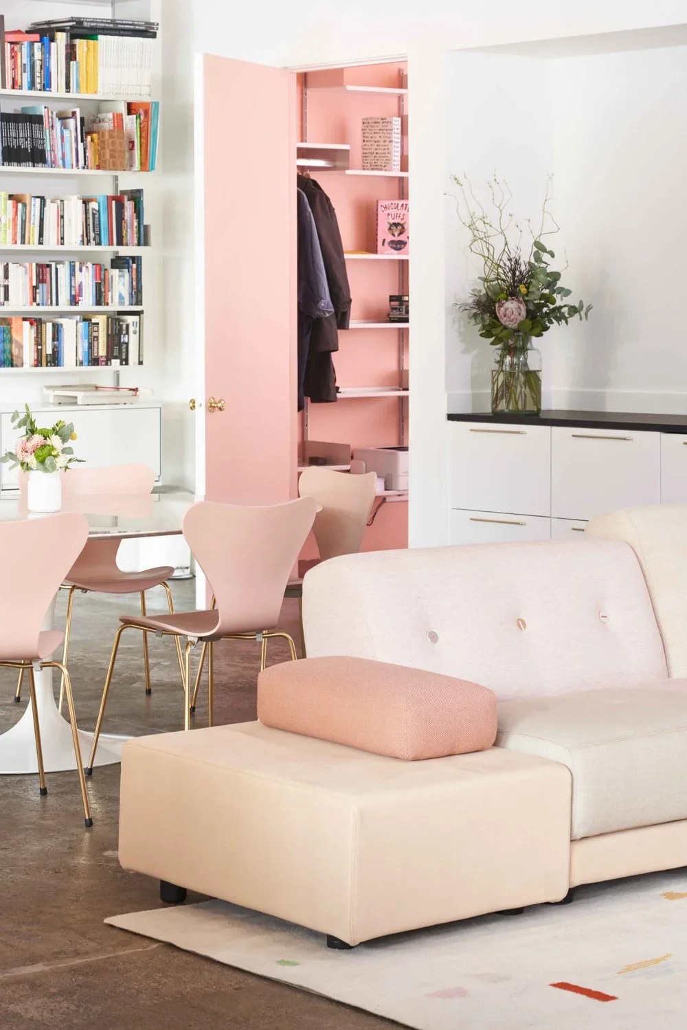 A cozy living space featuring a white sectional sofa with a pink cushion, a round dining table with pink chairs, a white bookshelf filled with books, a built-in cabinet with a floral arrangement, and an open closet with clothes and shelves painted pink.
