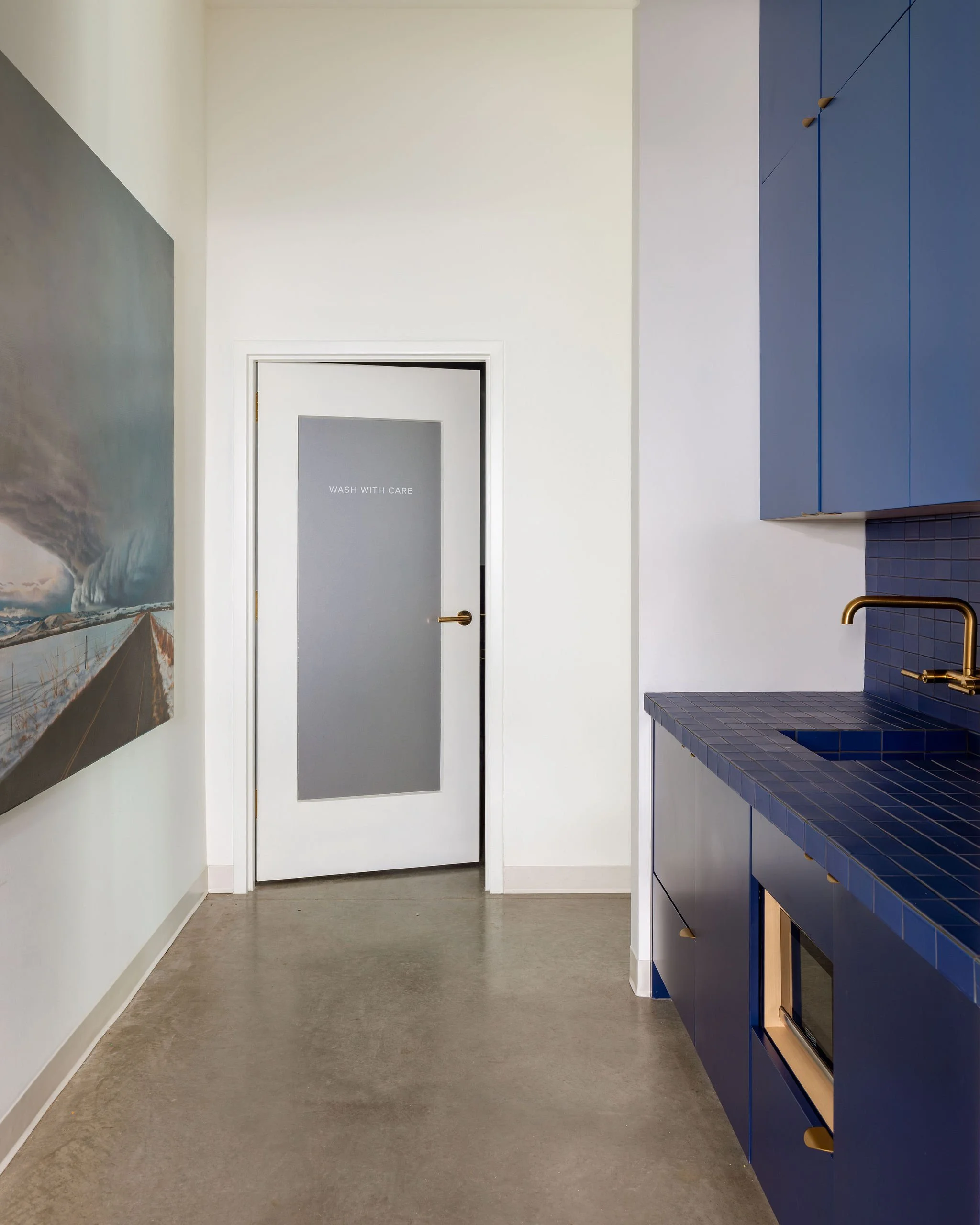 A modern kitchen with blue tiled countertop and cabinets, a gold faucet, concrete floor, and a door with frosted glass reading 'Wash with Care'.