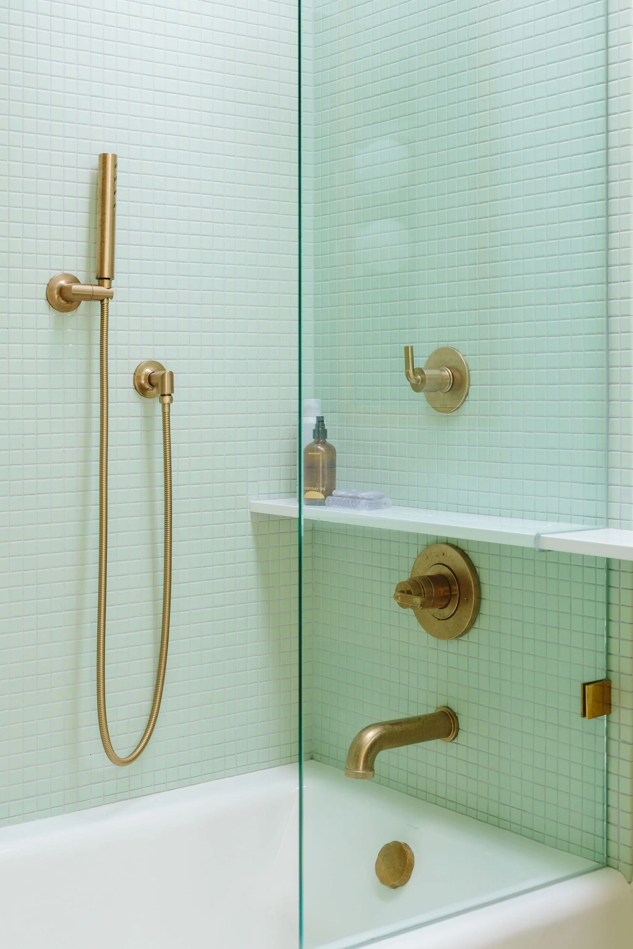 A modern shower with gold fixtures, green tiled walls, a built-in shelf with white towels and soap, and a glass partition.