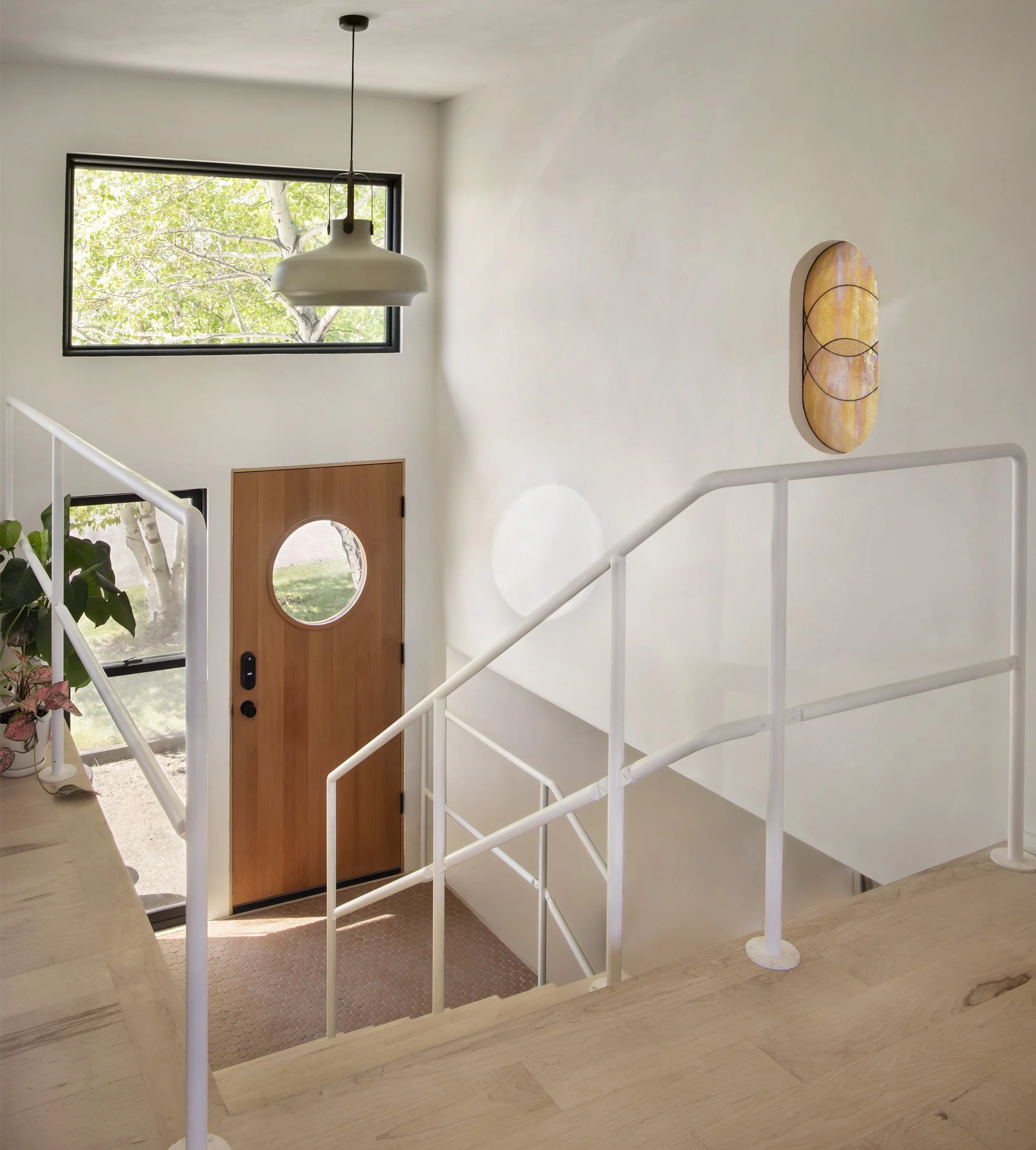 Interior view of a modern home entryway with a wooden front door with a circular window, a large rectangular window showing green trees outside, a white staircase with a metal railing, a hanging light fixture, and minimalist wall art.