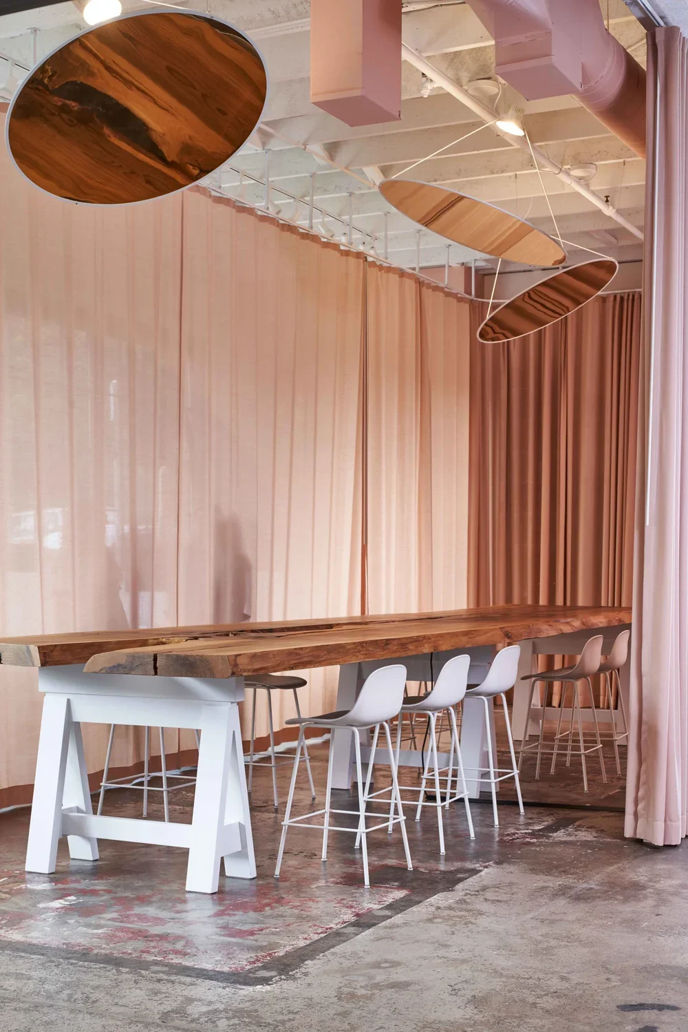 A modern interior space with a long wooden table, white and gray bar stools, pink curtains, and an overhead sculpture with three large, oval-shaped wooden panels suspended by thin wires.