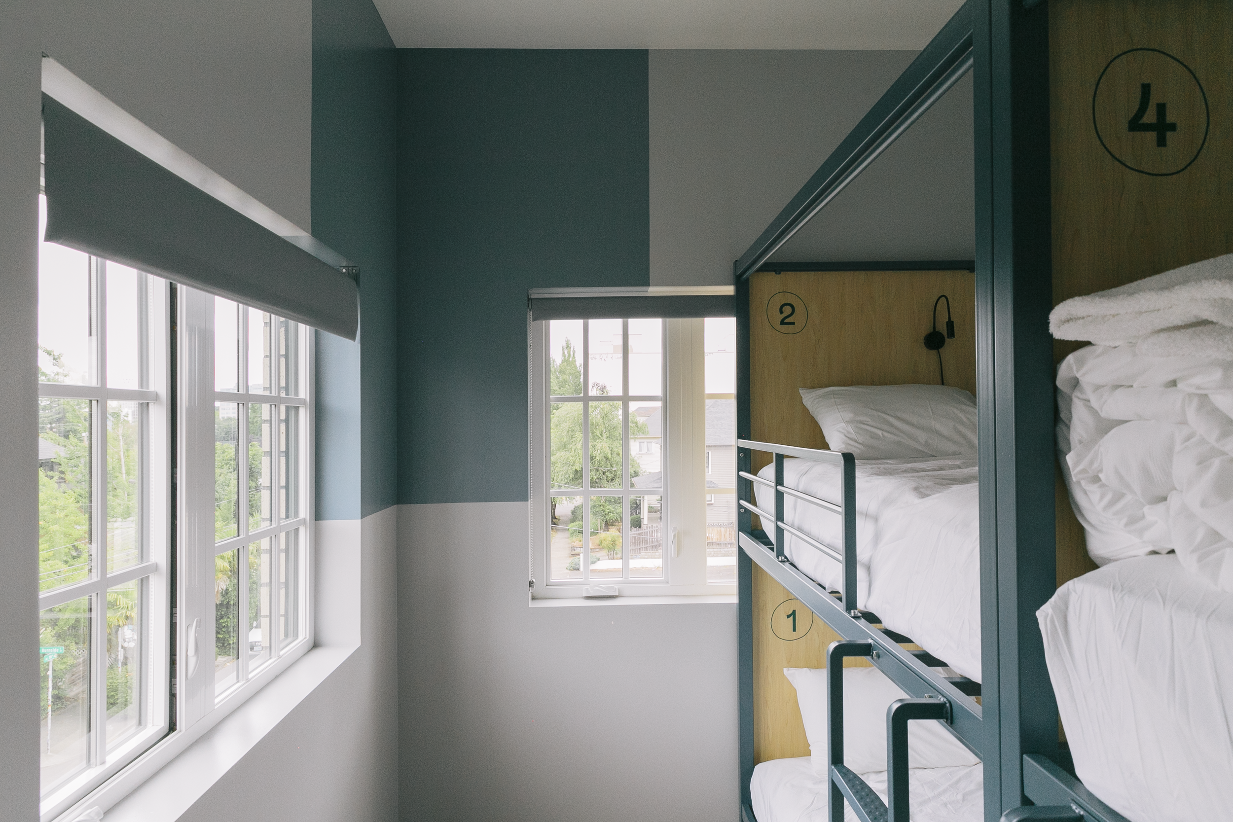 Room with bunk beds numbered 1 and 2, large windows with blinds, and a view of trees and houses outside.