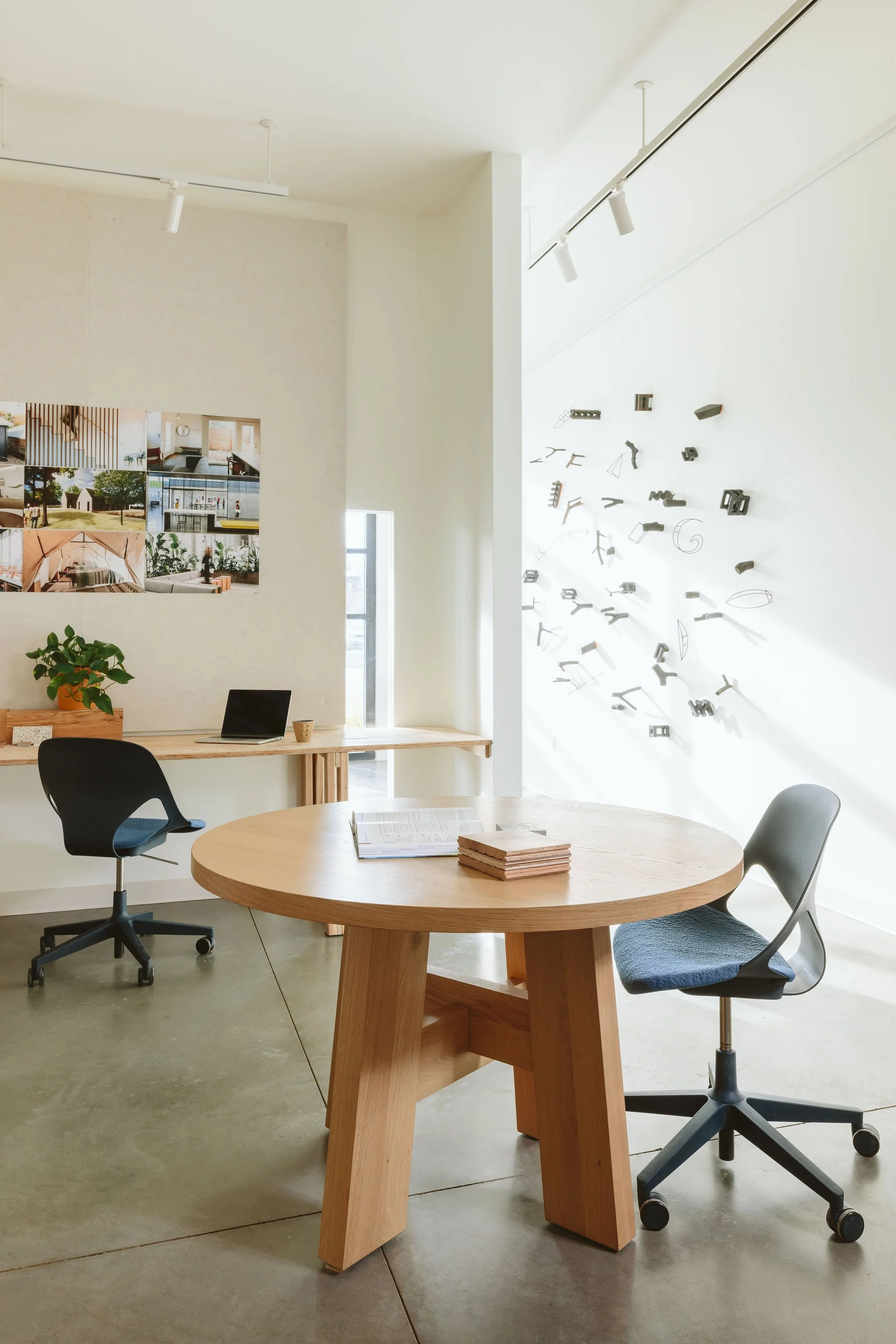 Modern office space with a round wooden table, two office chairs, a small desk with a plant, a laptop, books, and wall art including photos and hanging metal sculptures.