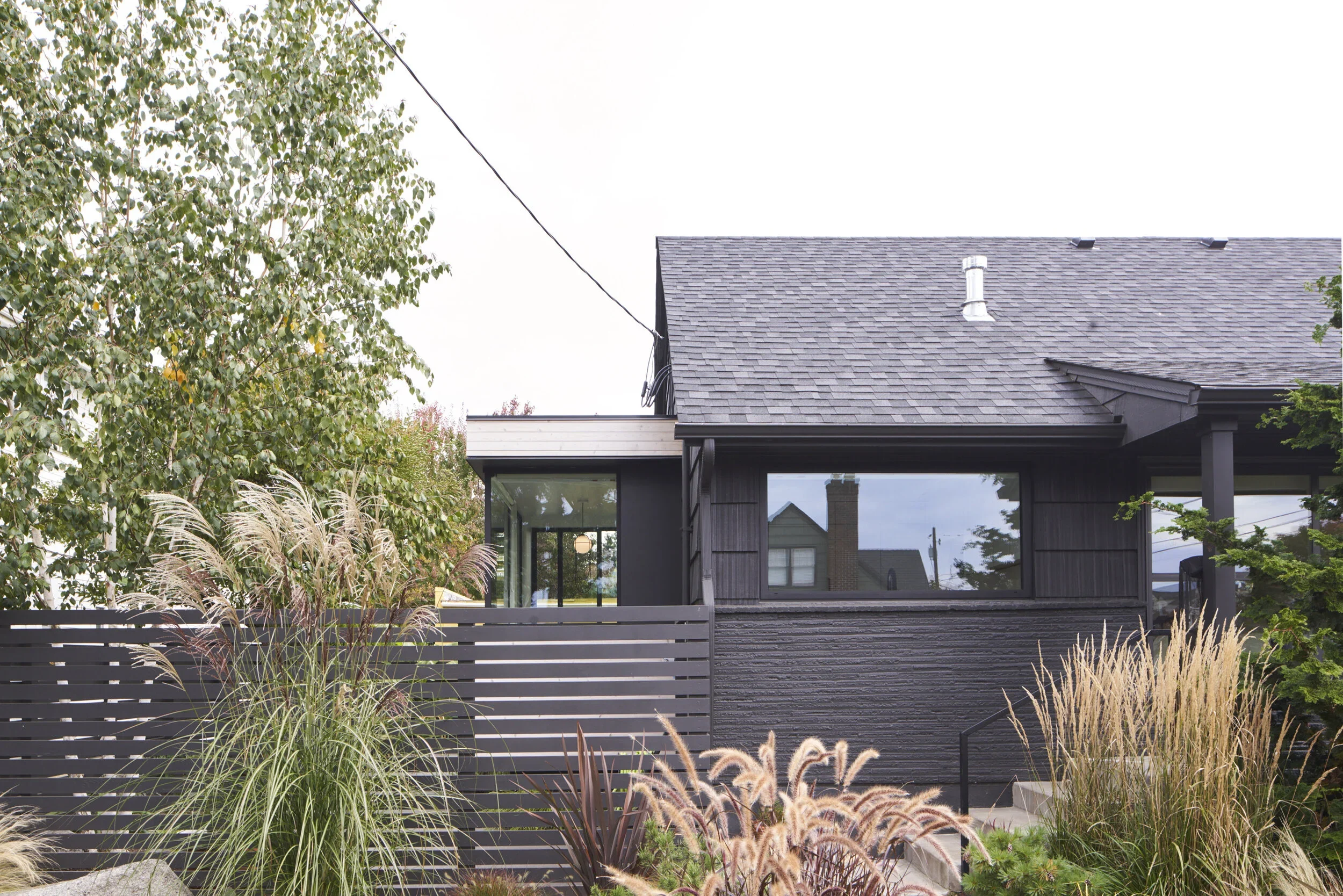 Modern black house with large rectangular window, sloped roof, surrounded by mature trees and ornamental grasses.