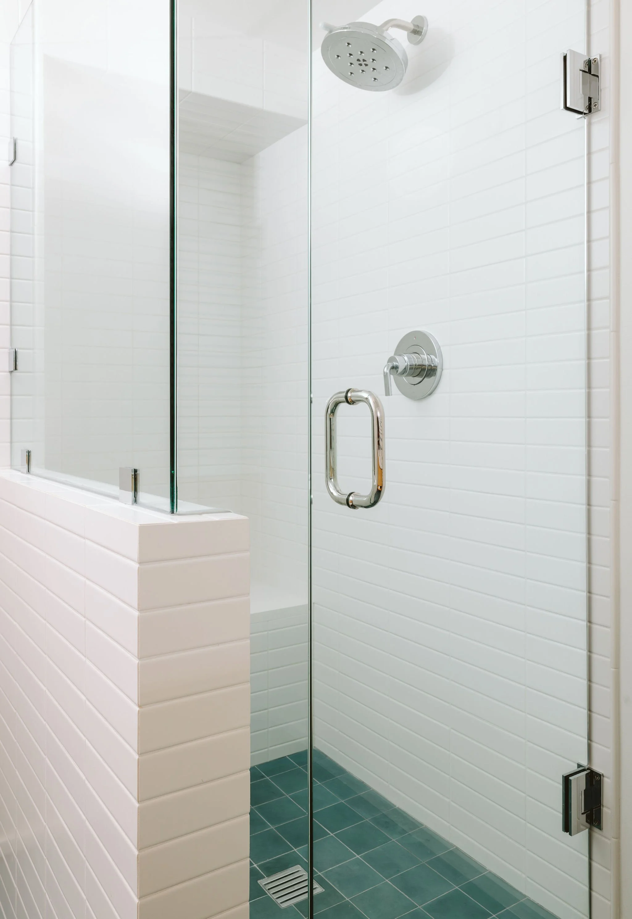 A modern, glass-enclosed shower with a rain showerhead, white tiled walls, a silver handle, and a drainage grate on the green tiled floor.