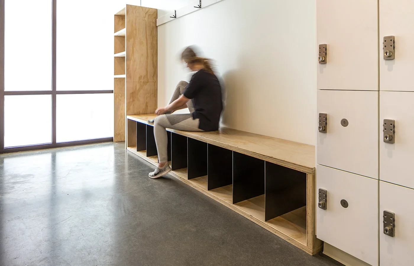 A woman sitting on a wooden and black cubby bench in a modern, minimalist locker area with natural light coming through large windows, metal storage lockers to the right, and a concrete floor.