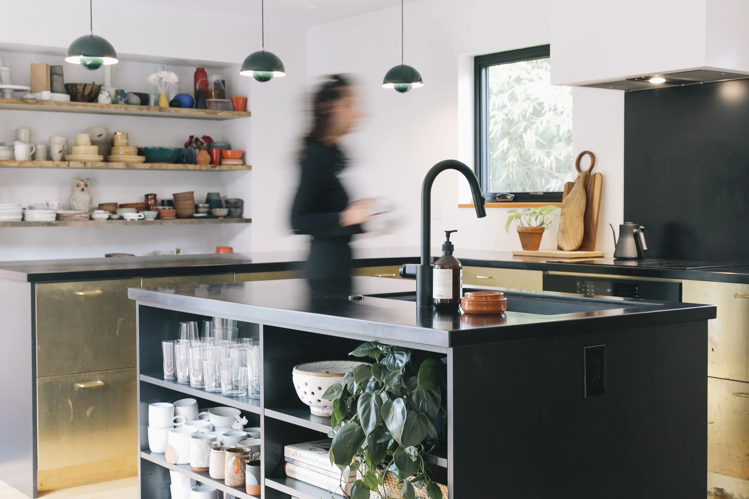 A woman in a black dress walking past a black kitchen island with glasses, a potted plant, and utensils, in a modern kitchen with open shelves filled with colorful dishes and bowls, and a window with greenery outside.