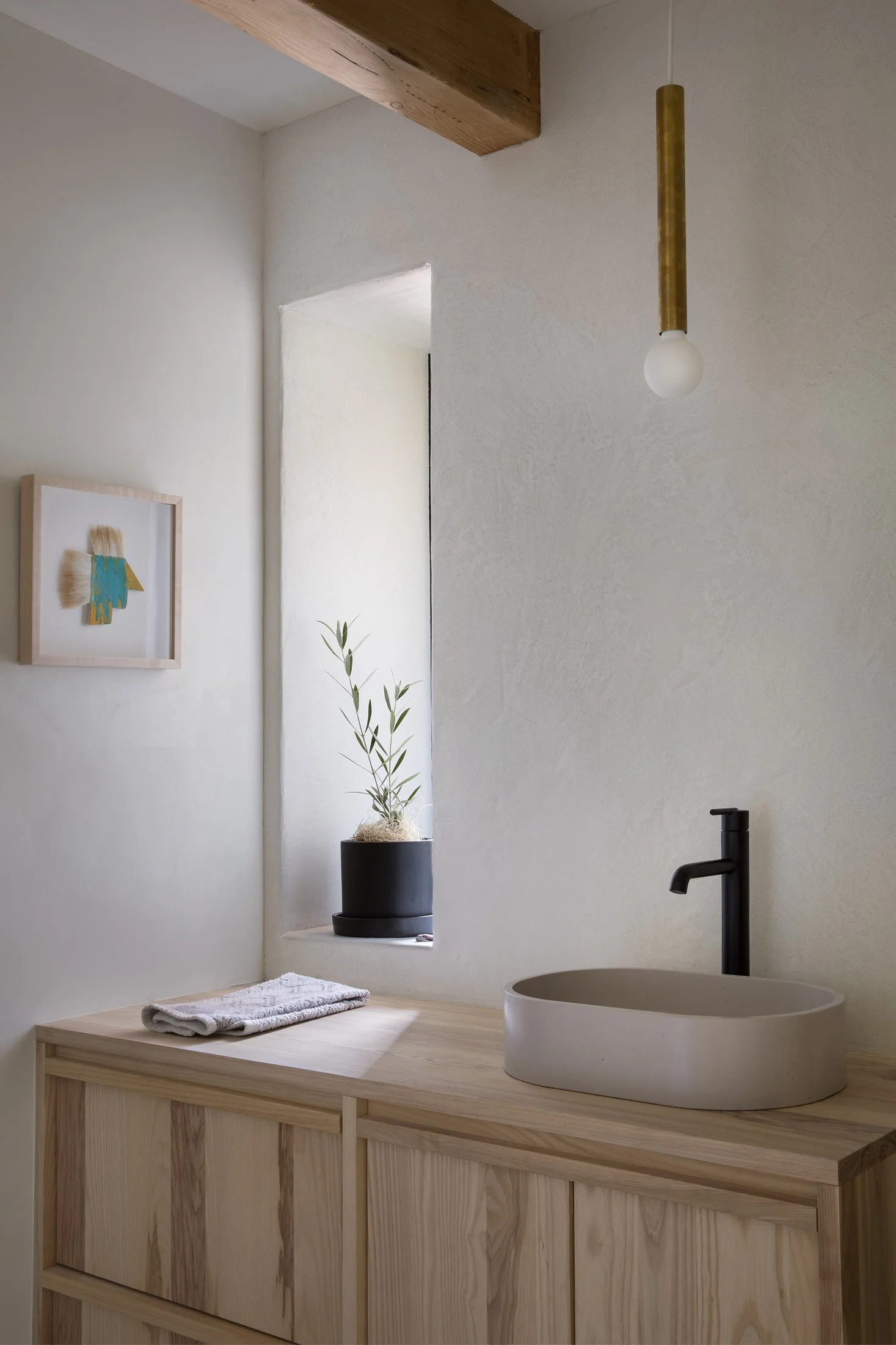 Minimalist bathroom with a wooden vanity, white oval sink, black faucet, small potted plant, framed artwork, towel, and hanging light fixture.