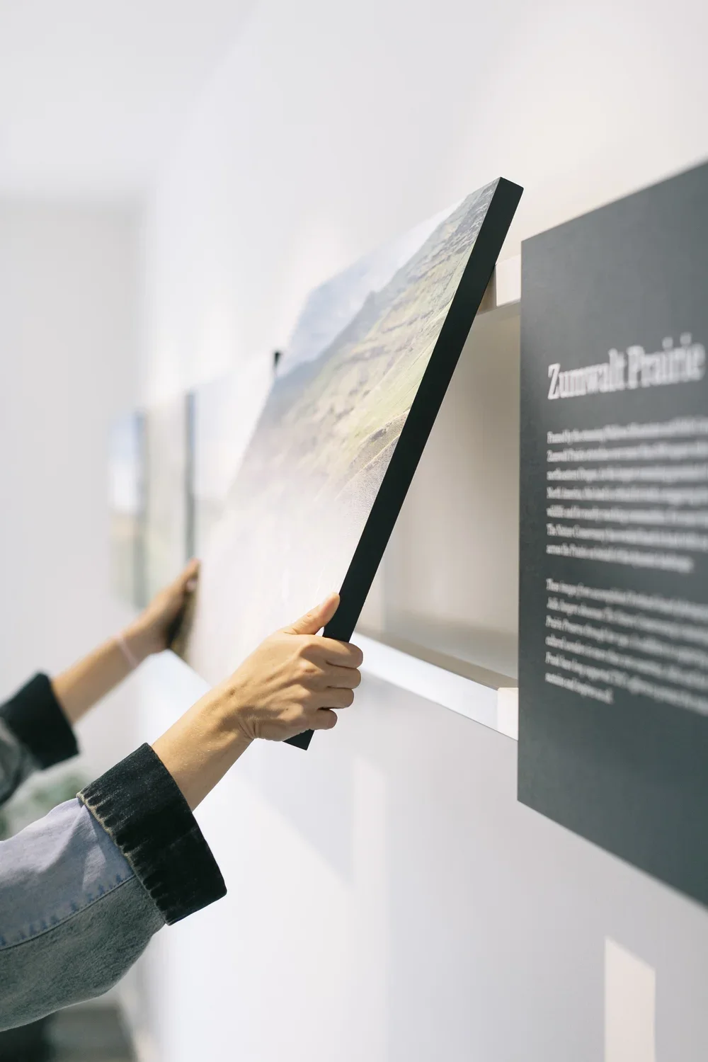 Person holding a landscape photograph at a gallery exhibit, with another displayed on the wall to the left.
