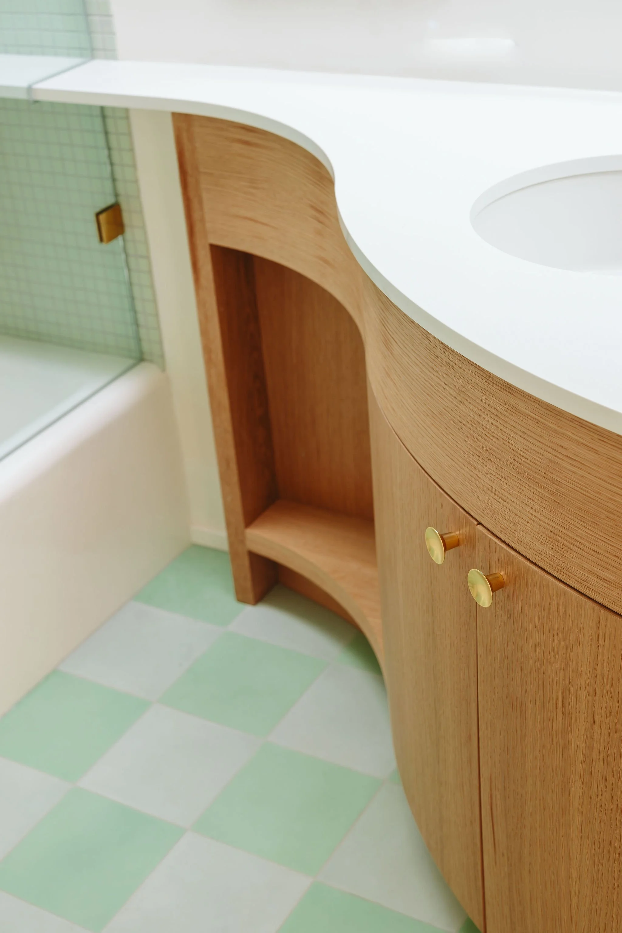Bathroom vanity with a white countertop, wooden cabinet doors with gold knobs, and a small open shelf. The floor has green and white checkered tiles.