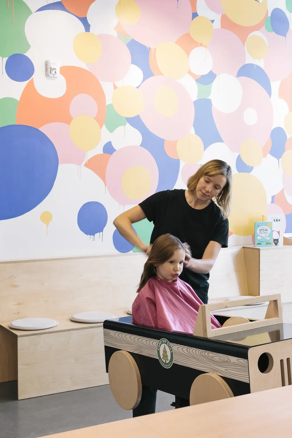 A woman with short blonde hair gives a haircut to a young girl with brown hair sitting in a wooden toy car, with a colorful balloon-patterned mural on the wall behind them.