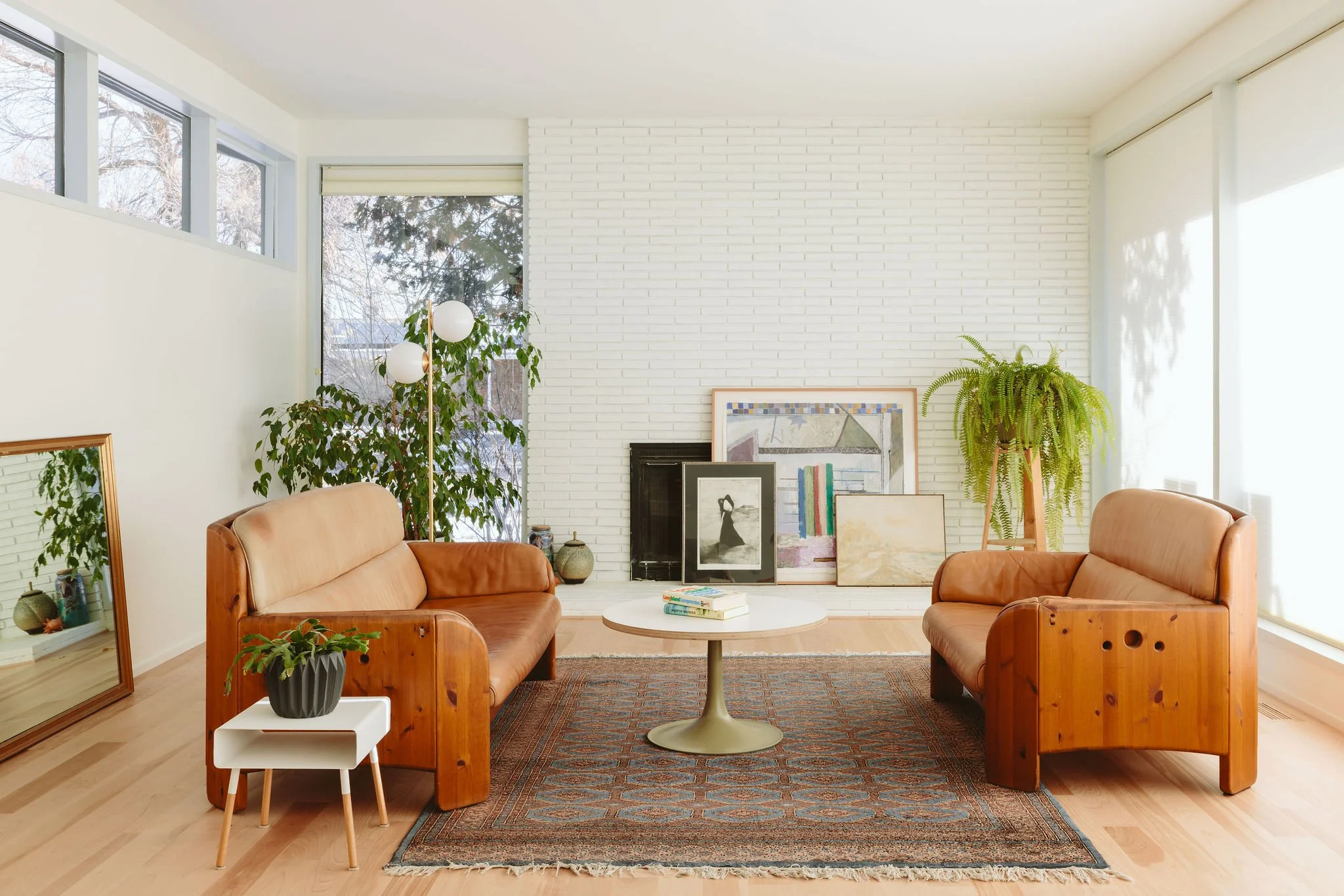 Sunlit living room with two leather sofas facing each other, a round white coffee table, a large mirror on the floor leaning against the wall, artwork and framed pictures on the white brick fireplace, and tall plants near windows with sheer white blinds.