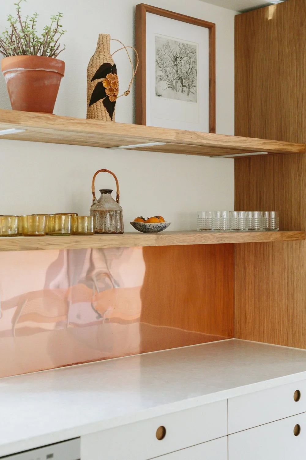 Open wooden shelf with decorative items including a potted plant, woven vase, vintage kettle, small bowl of oranges, and glasses, with framed art on the wall behind.