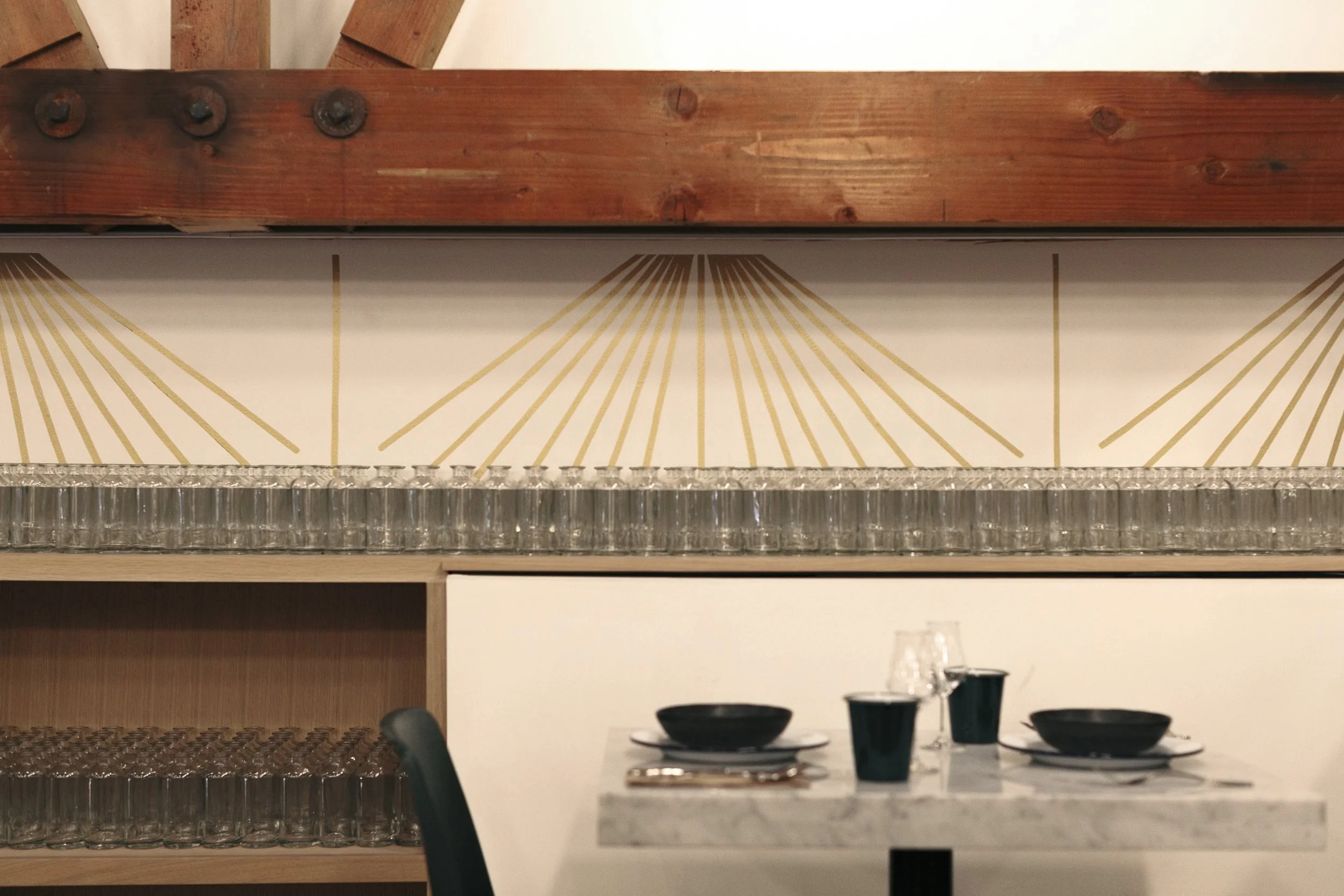 A dining table set with black bowls, black cups, and clear glasses, with a wood and glass shelf behind it holding additional glasses, and a decorative wall art with gold geometric lines in the background.