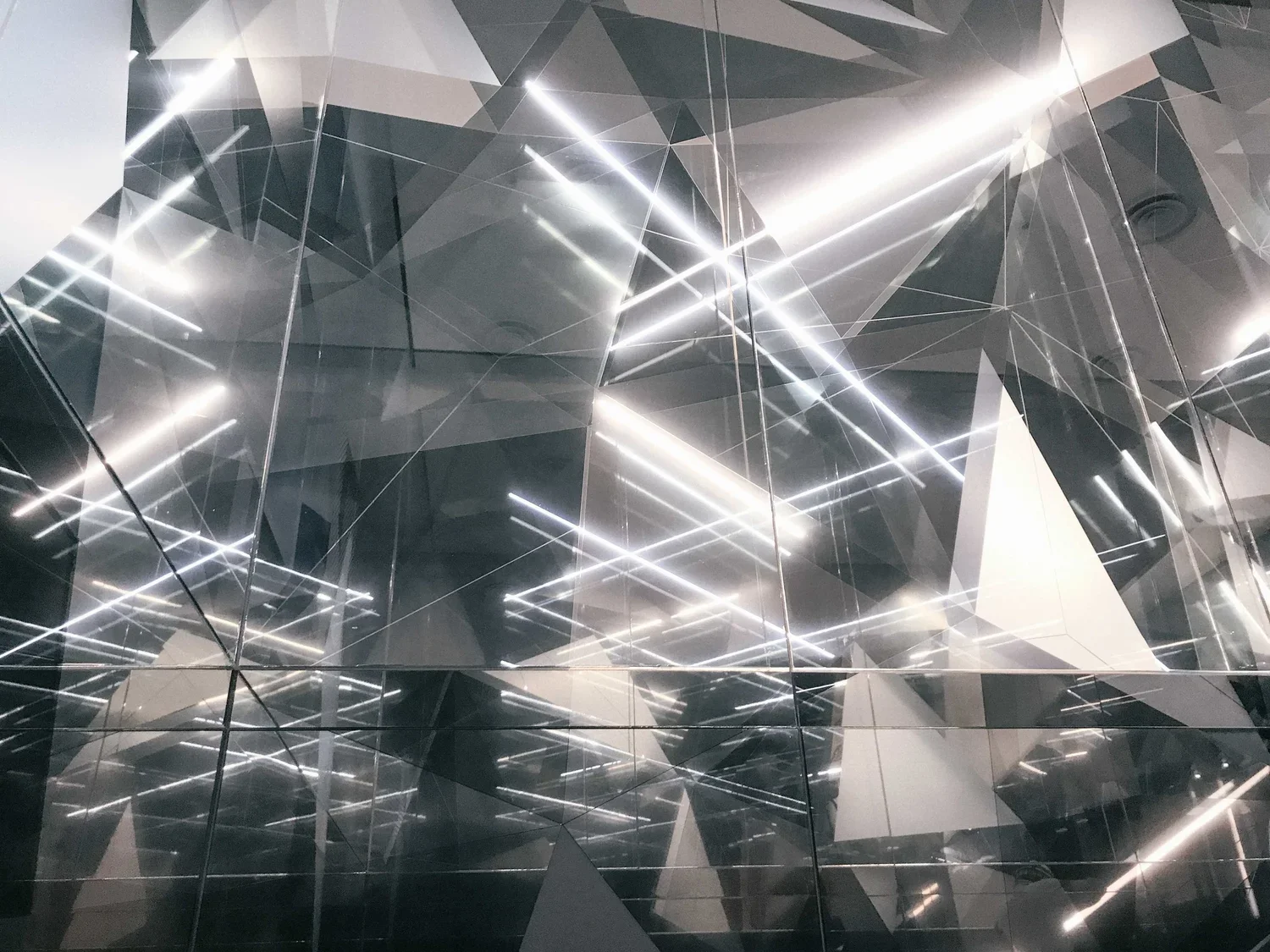 Reflection and abstract geometric patterns on a glass wall with bright linear lights and varying shades of gray and black.