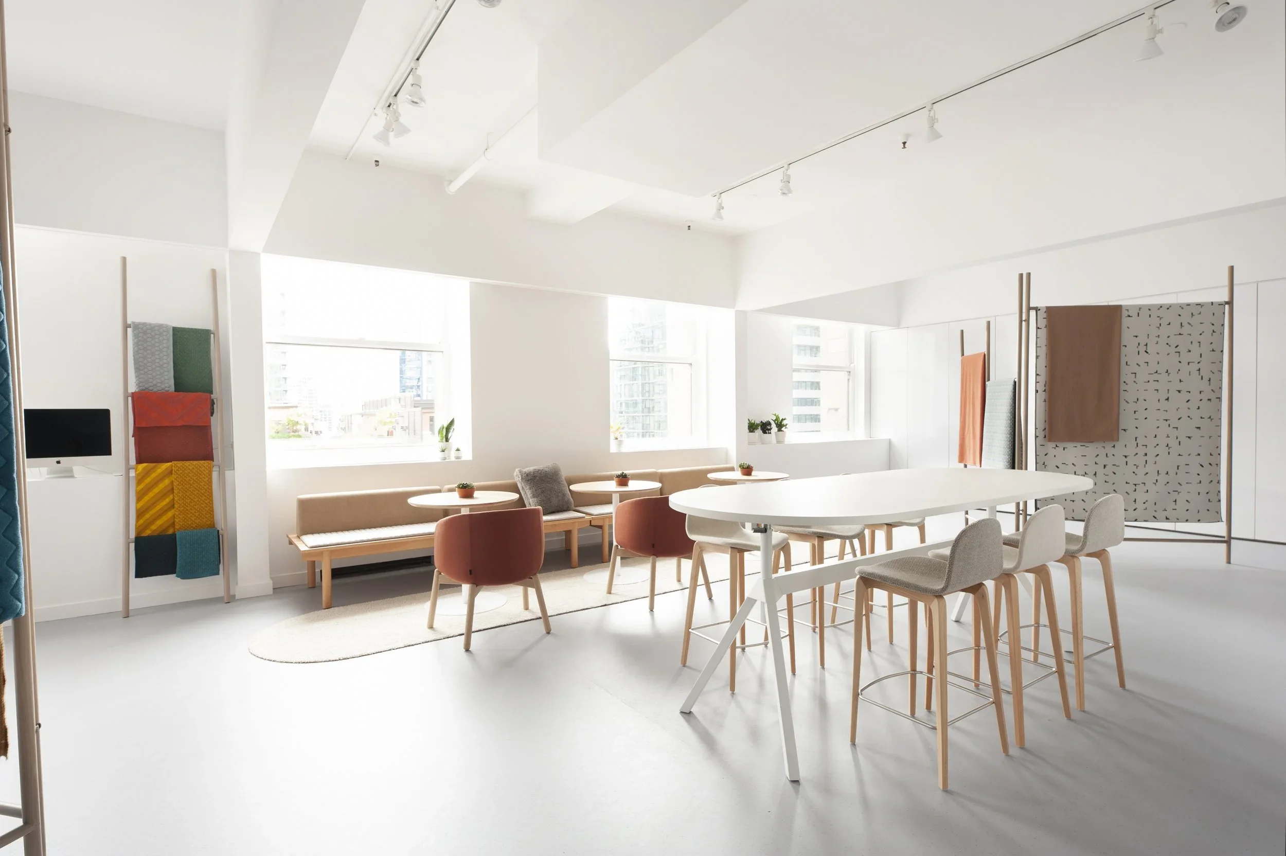Bright, modern, and minimalist interior space with white walls and large windows, featuring a long white counter with chairs, small tables with plants, and fabric panels on display