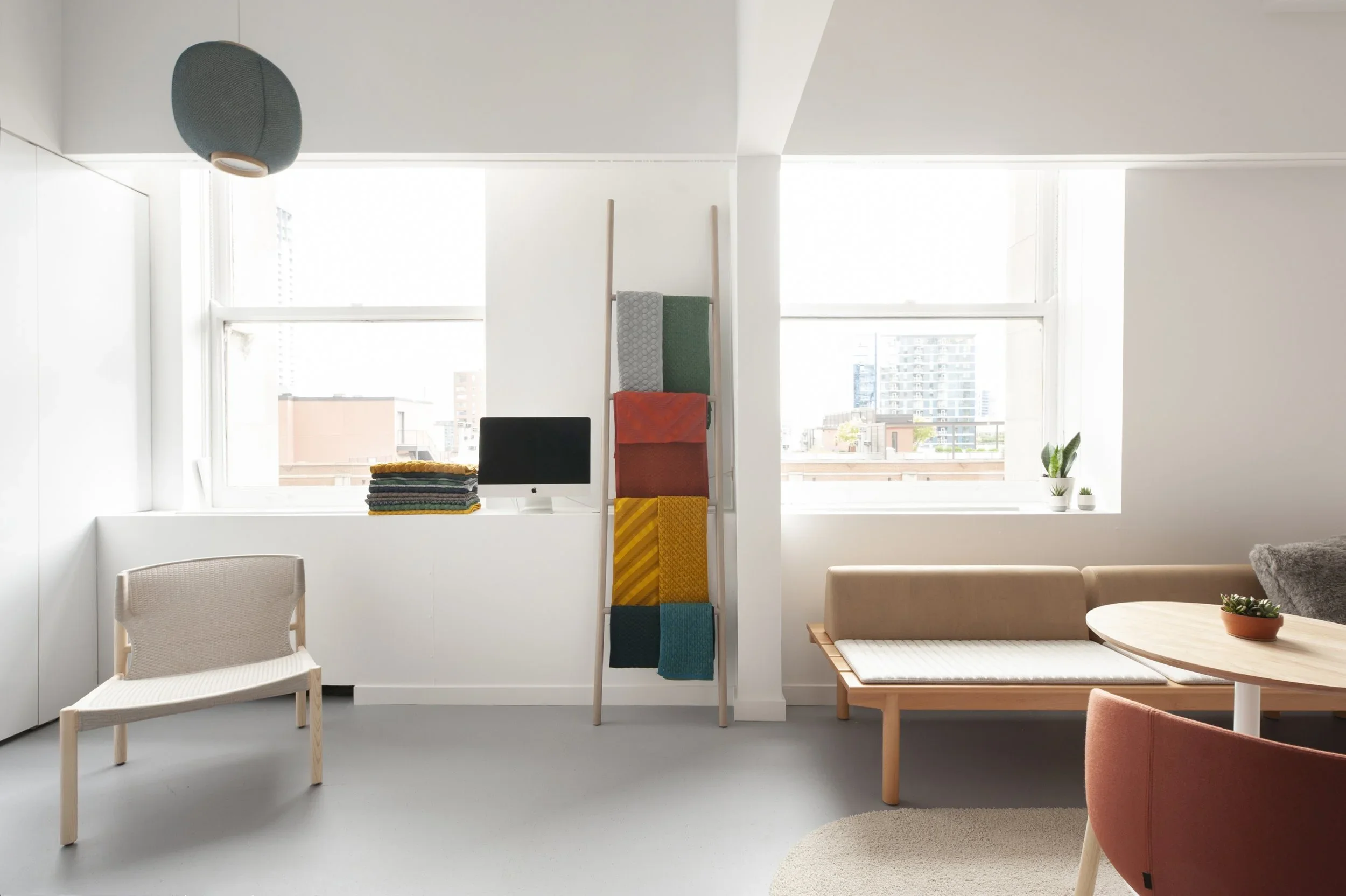 Modern living room with large windows, beige couch, round wooden table, white chair, colorful towel ladder, stacked towels, potted plants, and a blue hanging lamp.