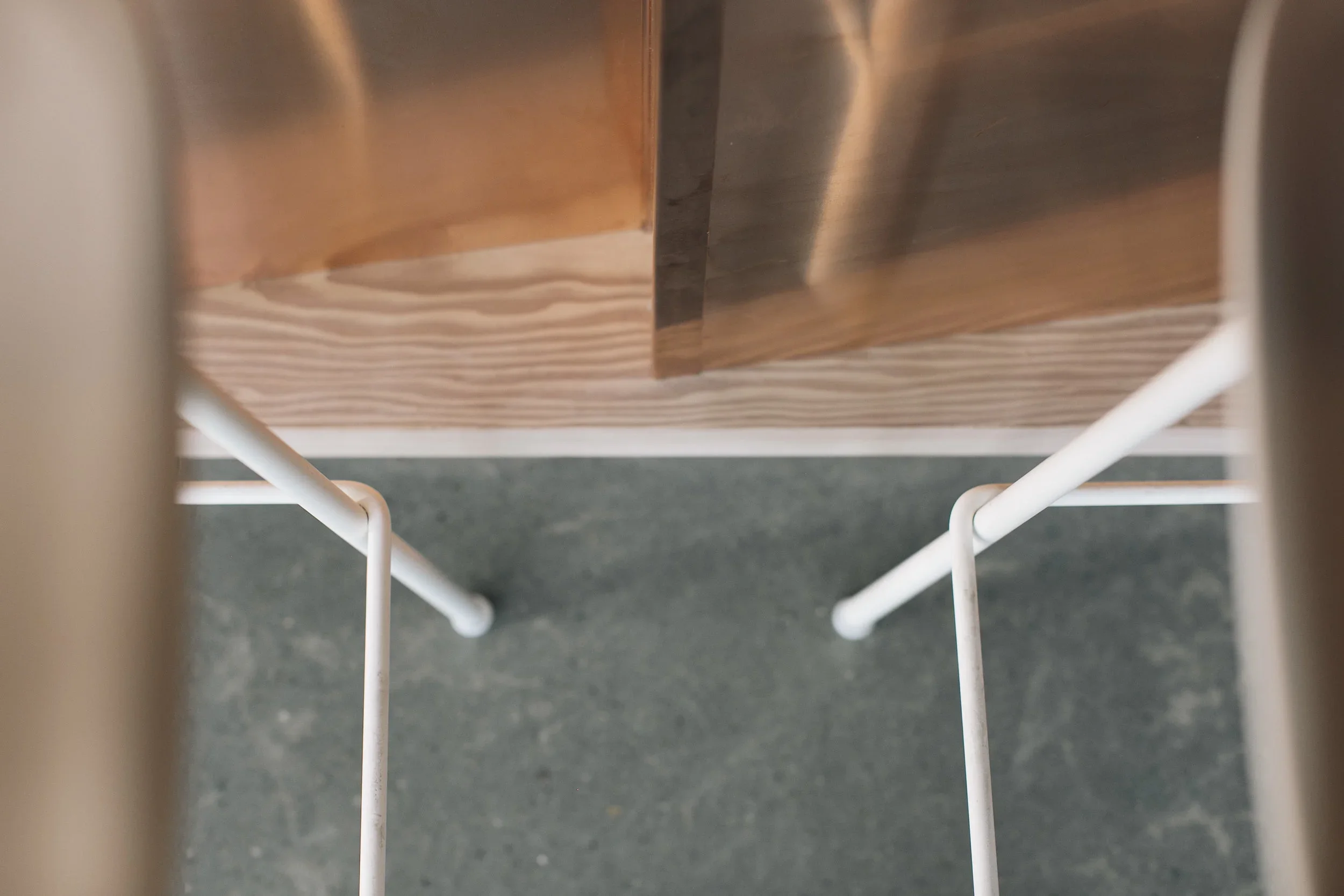 Looking down under a wooden table, two white metal chair legs are visible on dark flooring.