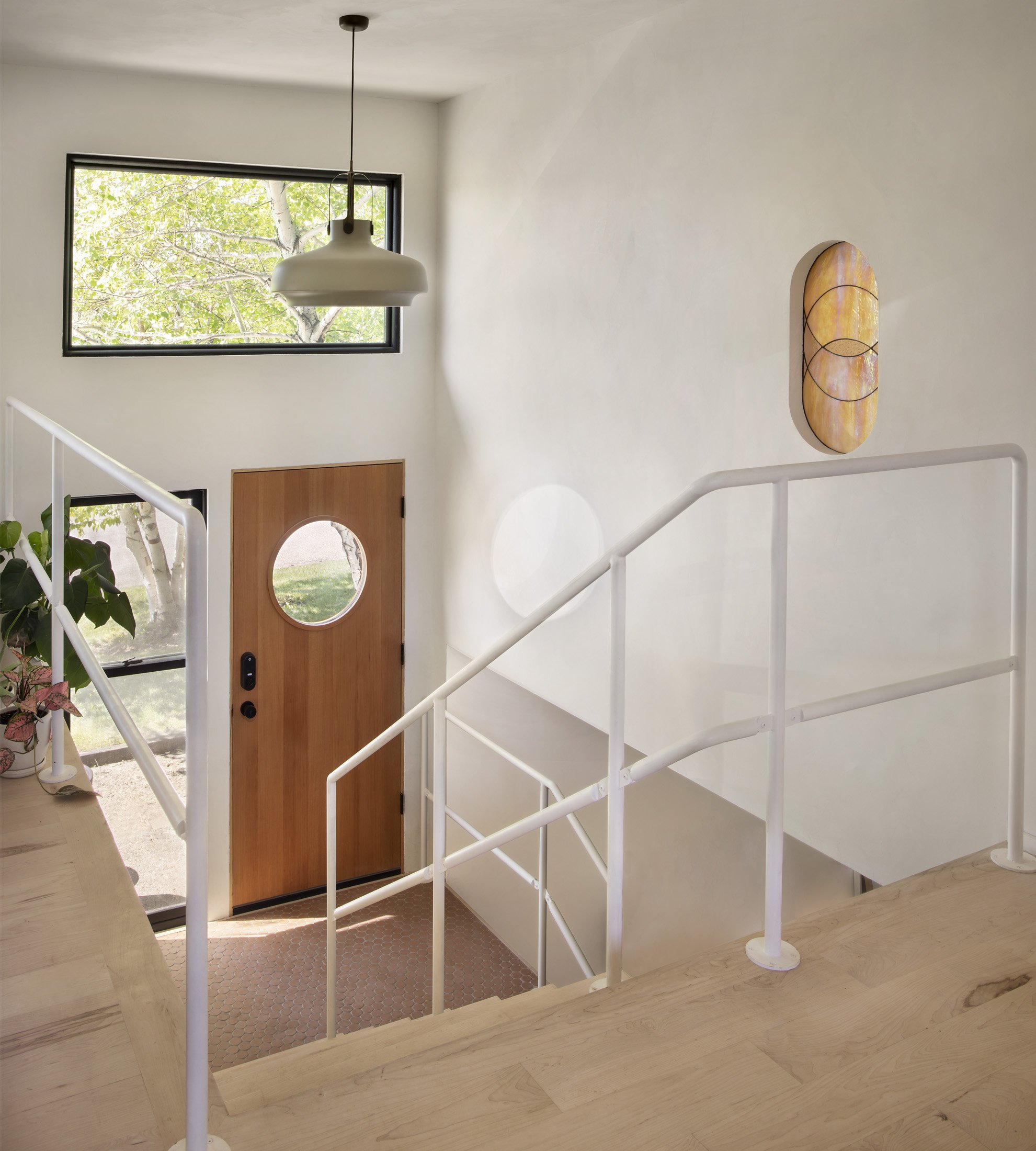 Entryway of a modern home with a wooden door featuring a round window, a large rectangular window above, and a staircase with white railing. A hanging ceiling light and abstract wall art are visible.