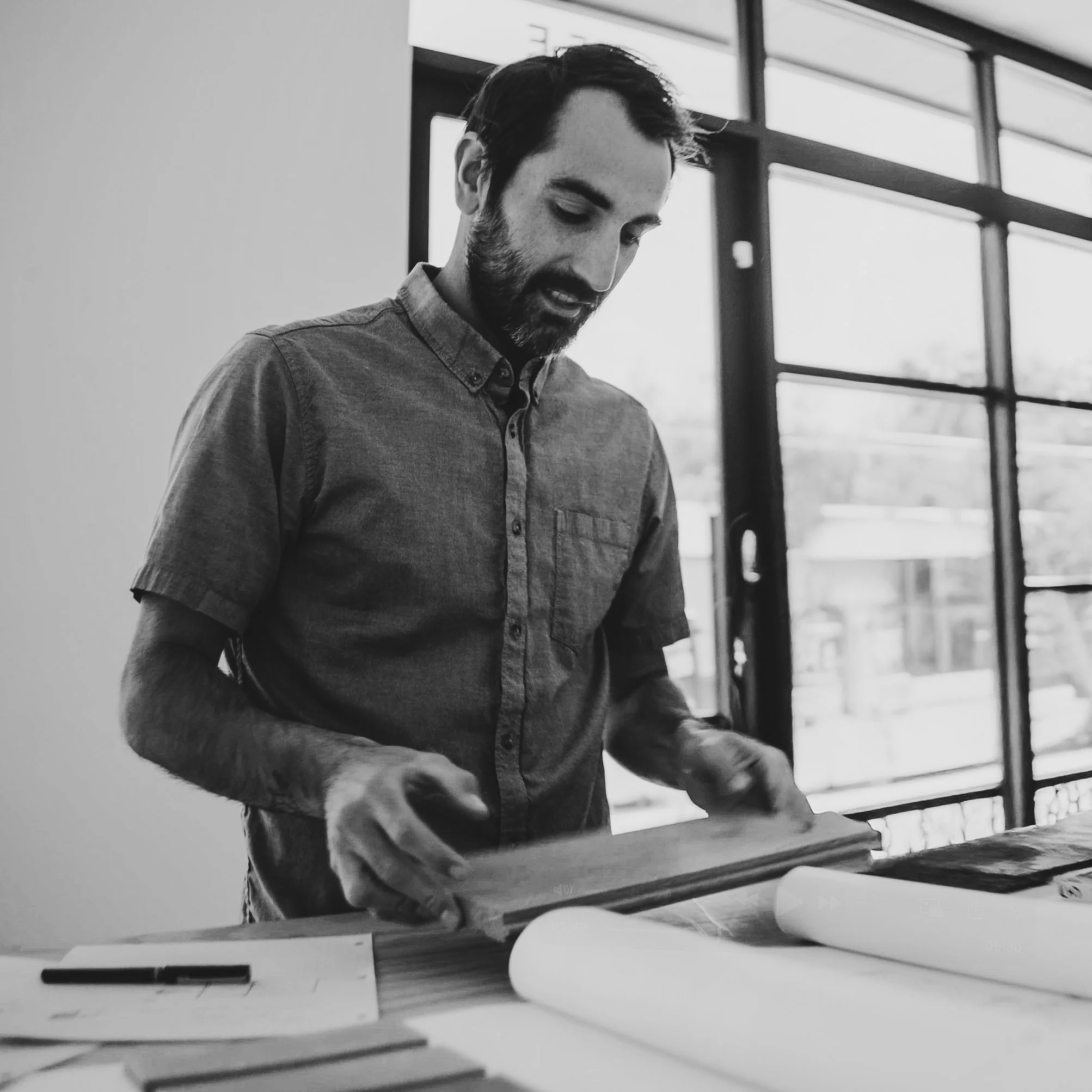 A man with dark hair and a beard looking down at a large book or blueprint on a table. There are rolled-up papers and a pen on the table as well. He is indoors near large windows.