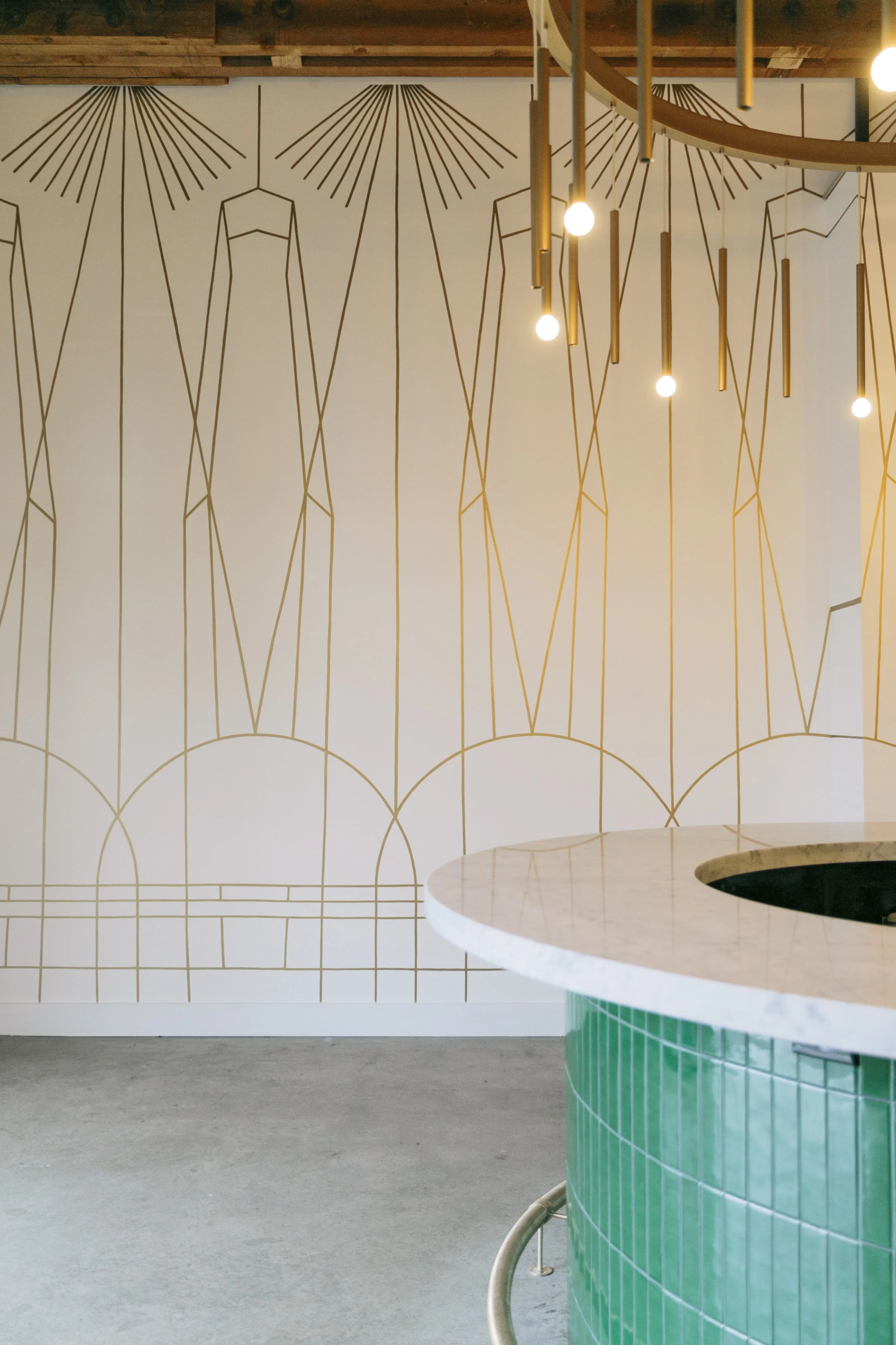 Interior of a modern space featuring a white wall with gold geometric line art and hanging gold and bulb pendant lights above a curved green tiled counter.