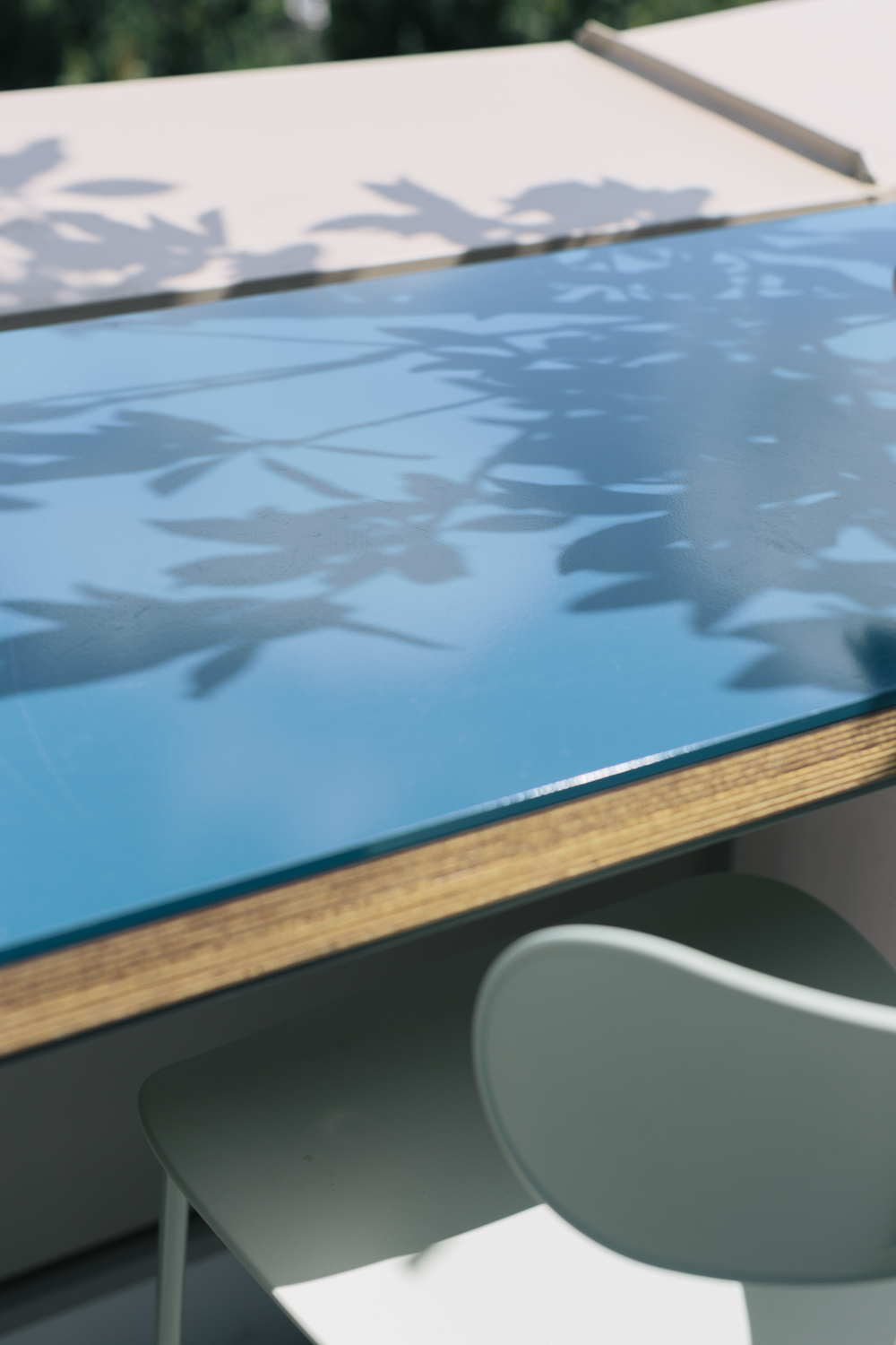 A table with a glass top, reflecting shadows of leaves and a chair in front of it.