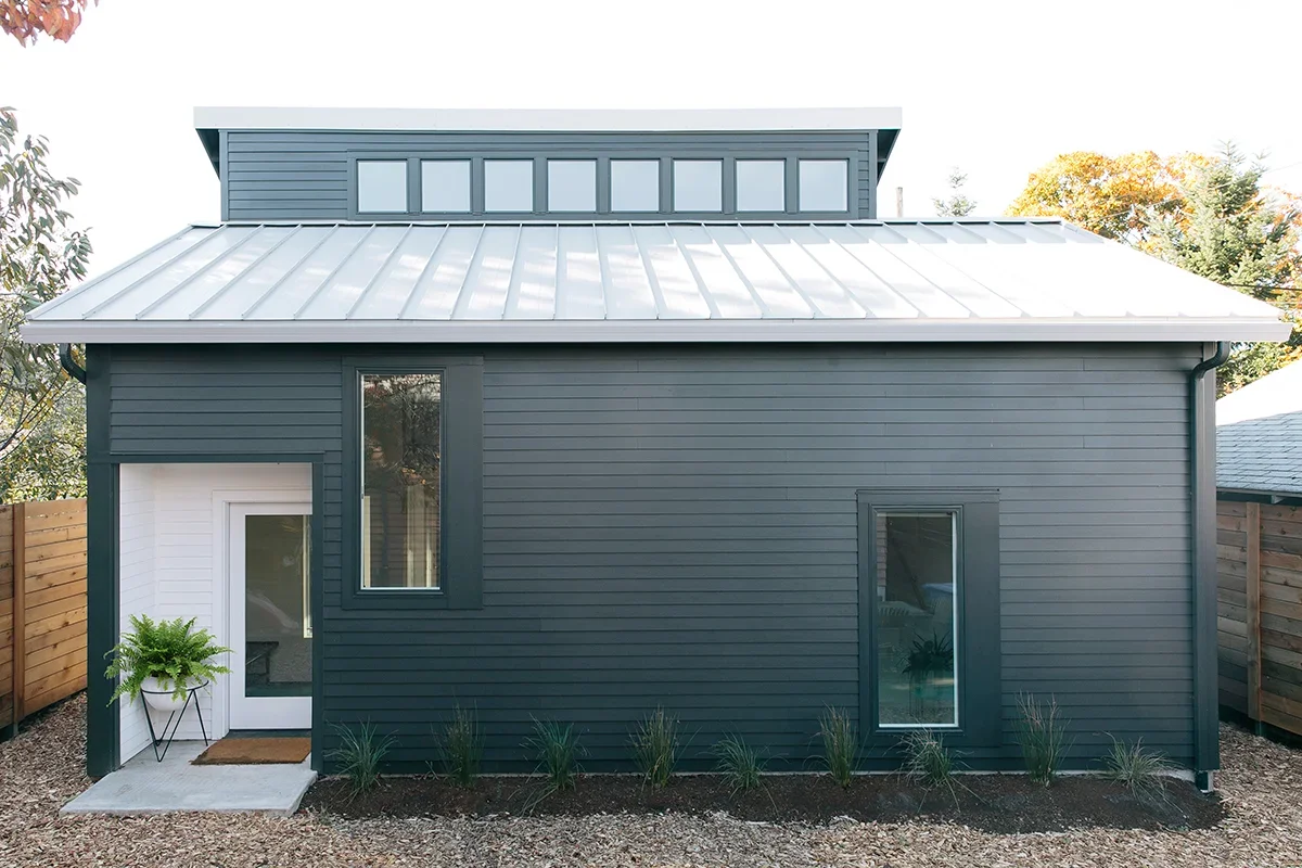 Modern dark gray tiny house with a metal roof, multiple windows, and a small front porch with a white door and a potted plant.