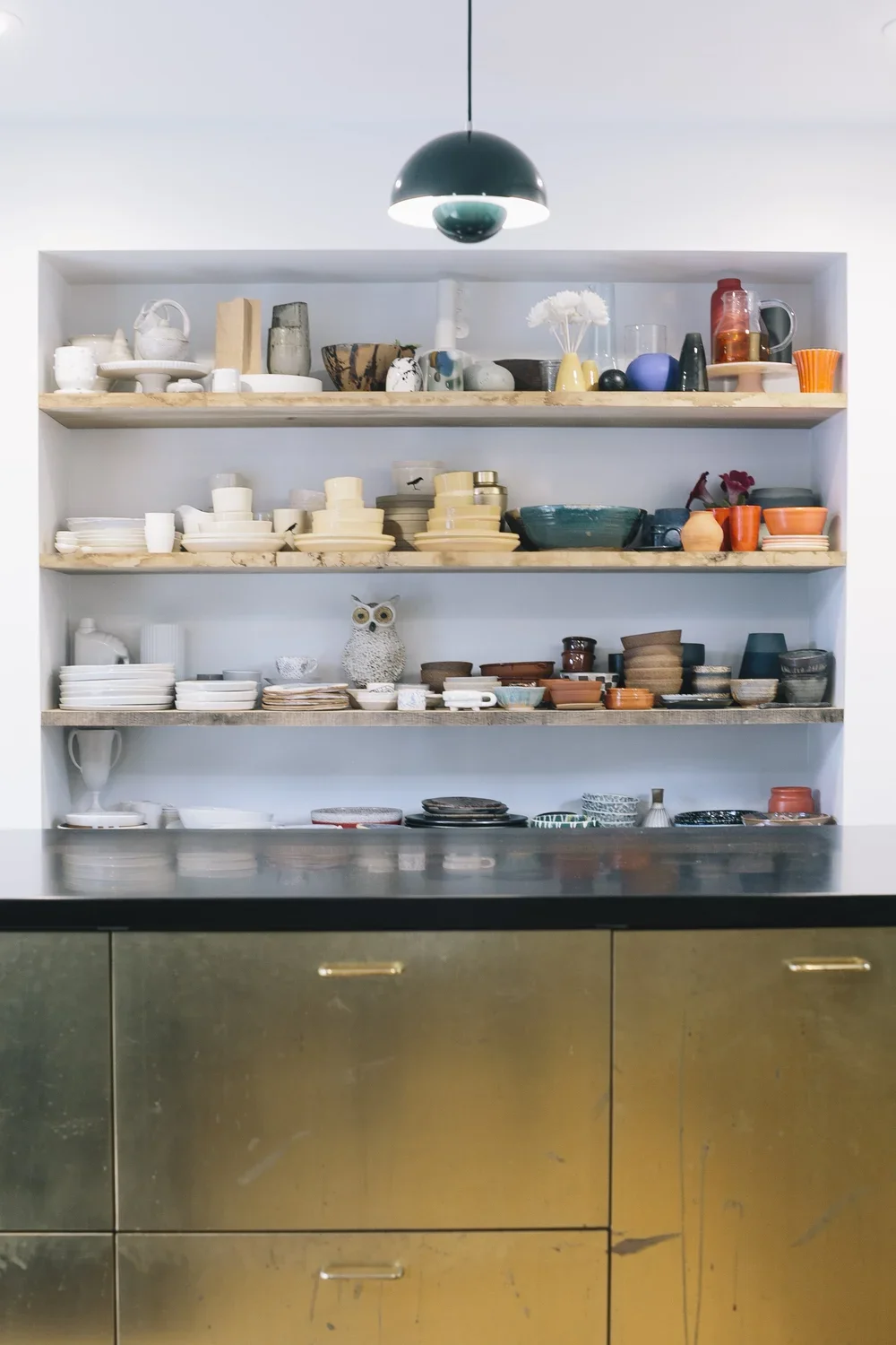 Open shelves filled with various kitchenware including bowls, plates, cups, and decorative items, with a dark countertop and a gold metallic cabinet below, and a green hanging pendant light overhead.